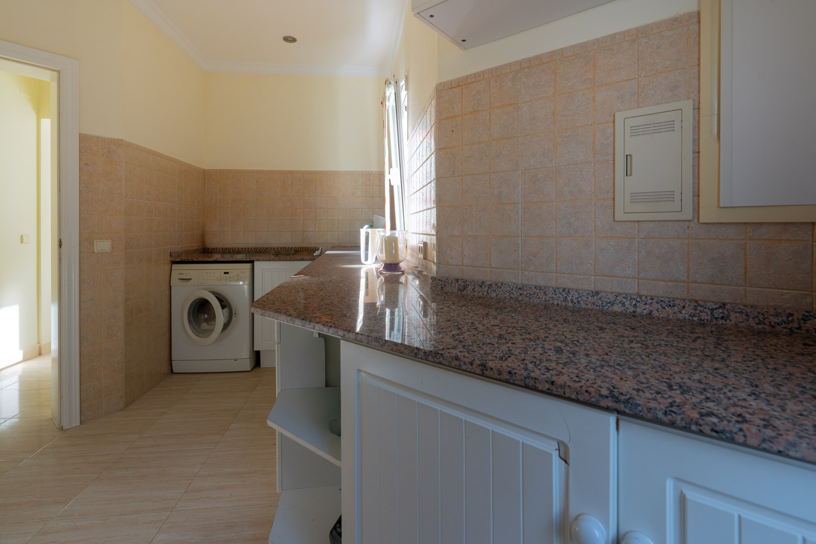 Lavadero práctico con encimera de granito, armarios blancos y zona de lavado, iluminado por luz natural y revestido con azulejos cerámicos.