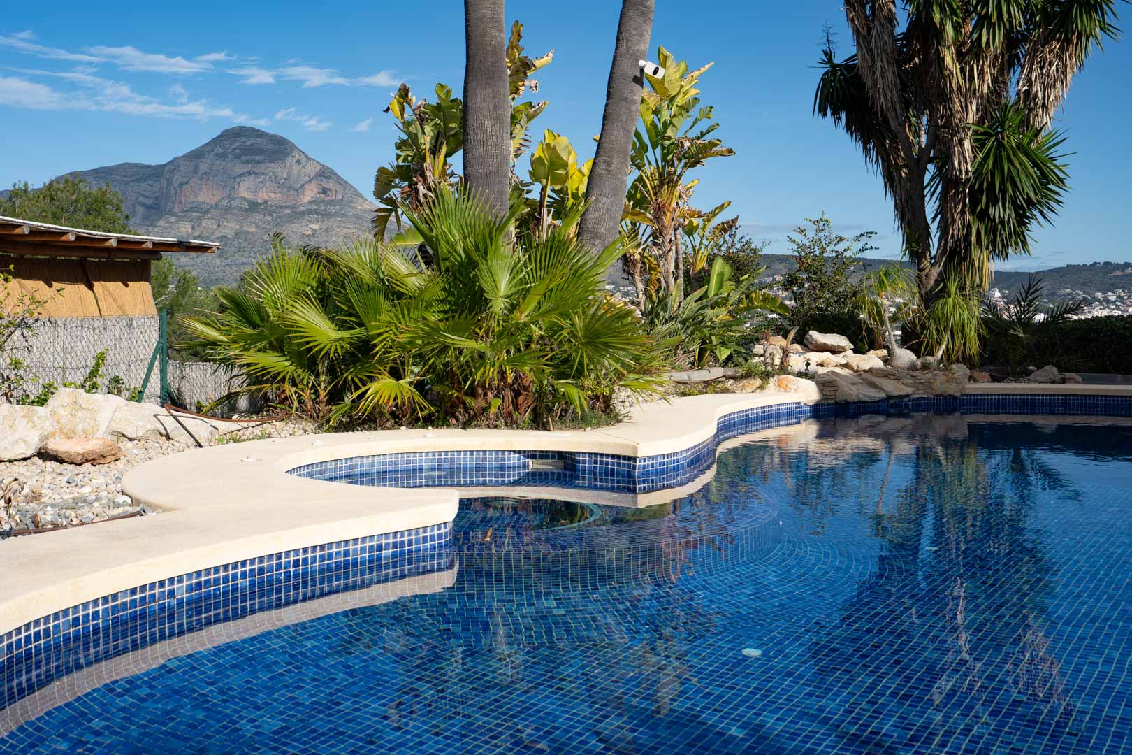Piscina de gresite azul con formas curvas y jacuzzi integrado, rodeada de vegetación tropical y con vistas frontales al macizo del Montgó.