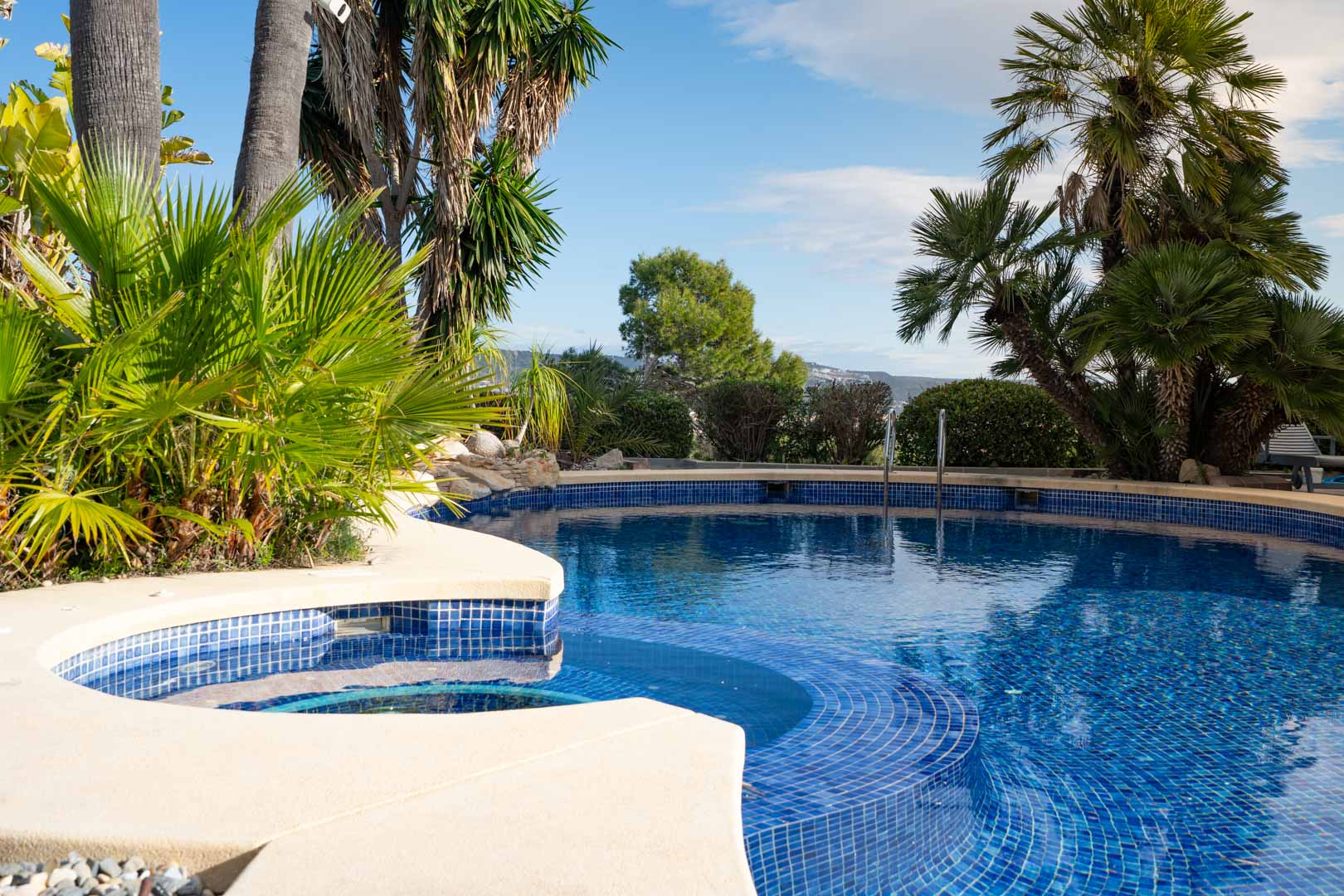 Piscina exterior de diseño curvo revestida con mosaico azul, integrada en un entorno de vegetación tropical y palmeras autóctonas.