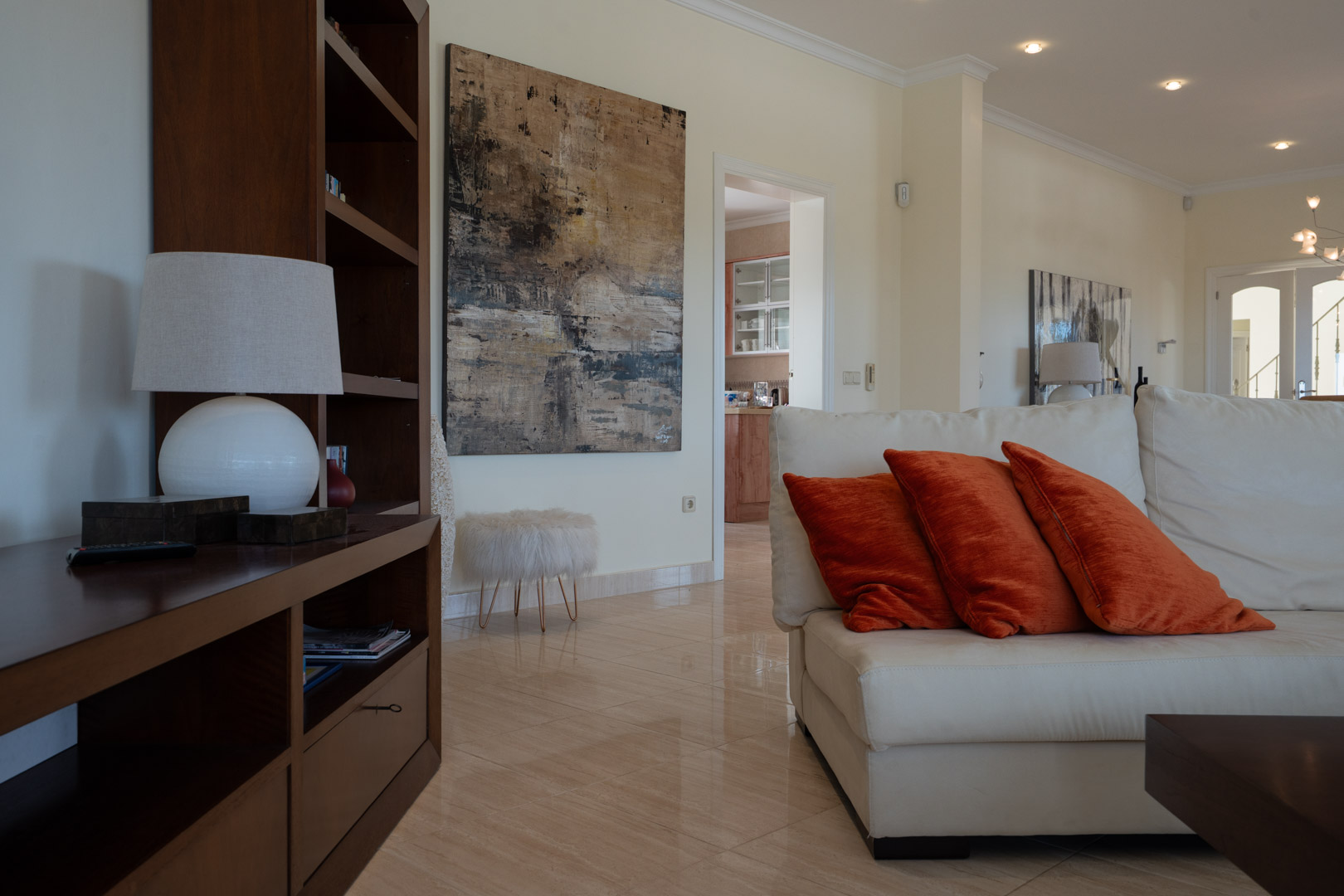 Salón con suelos de gres pulido, sofá de cuero blanco con cojines naranjas y estantería de madera oscura junto a una gran obra de arte abstracta.