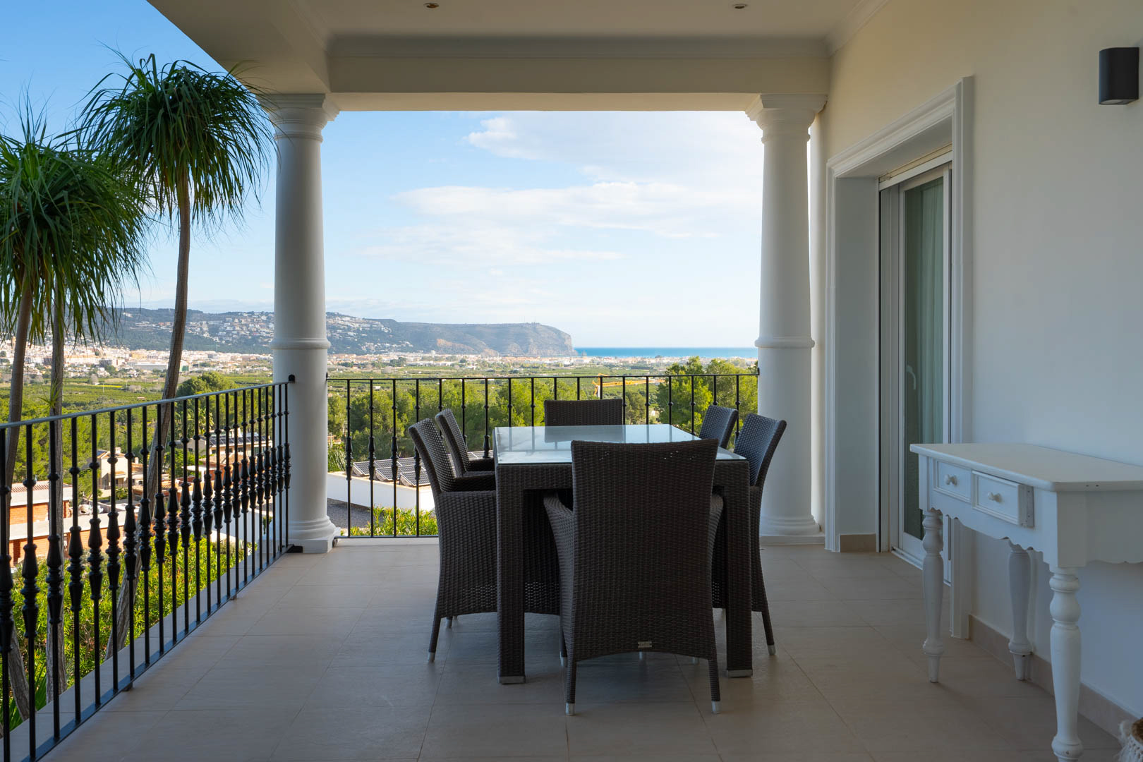 Terraza cubierta con columnas clásicas y barandilla de forja, equipada con comedor exterior y vistas panorámicas al mar Mediterráneo y al valle.