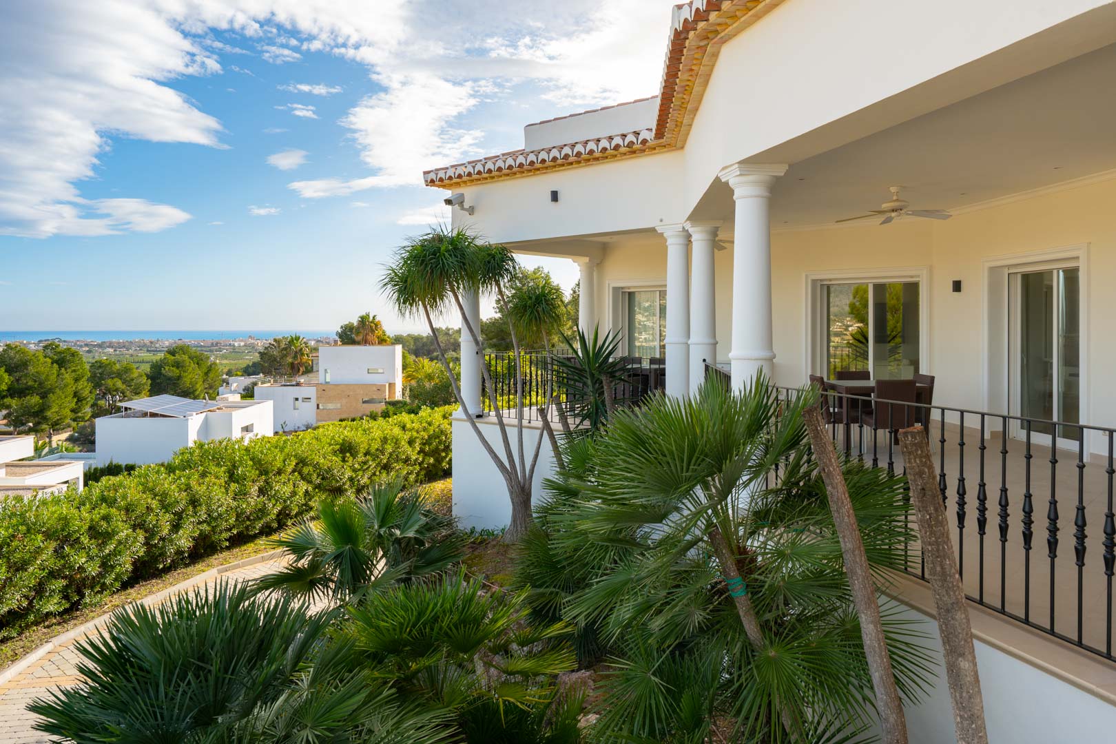 Terraza cubierta con columnas clásicas y barandilla de forja, rodeada de vegetación tropical y con vistas panorámicas al mar Mediterráneo y al entorno costero.