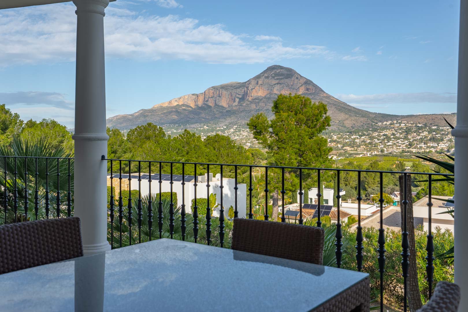 Terraza cubierta con mesa de comedor exterior y barandilla de hierro forjado, ofreciendo vistas panorámicas despejadas al macizo del Montgó.