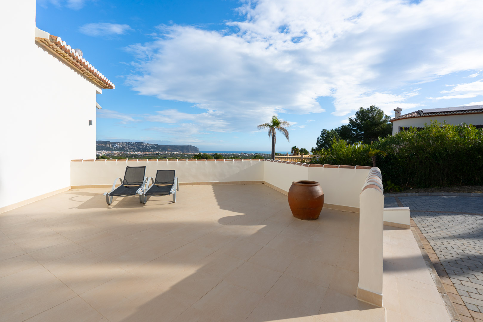 Terraza solárium con suelos cerámicos, tumbonas y vistas panorámicas al mar Mediterráneo y la costa, delimitada por un muro blanco con remate de teja.