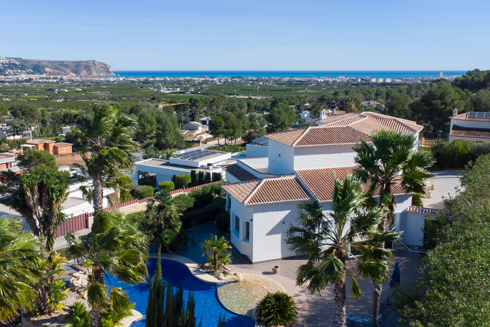 Vista aérea de la villa blanca con piscina azul, palmeras maduras y vistas panorámicas hacia el mar Mediterráneo y la costa.