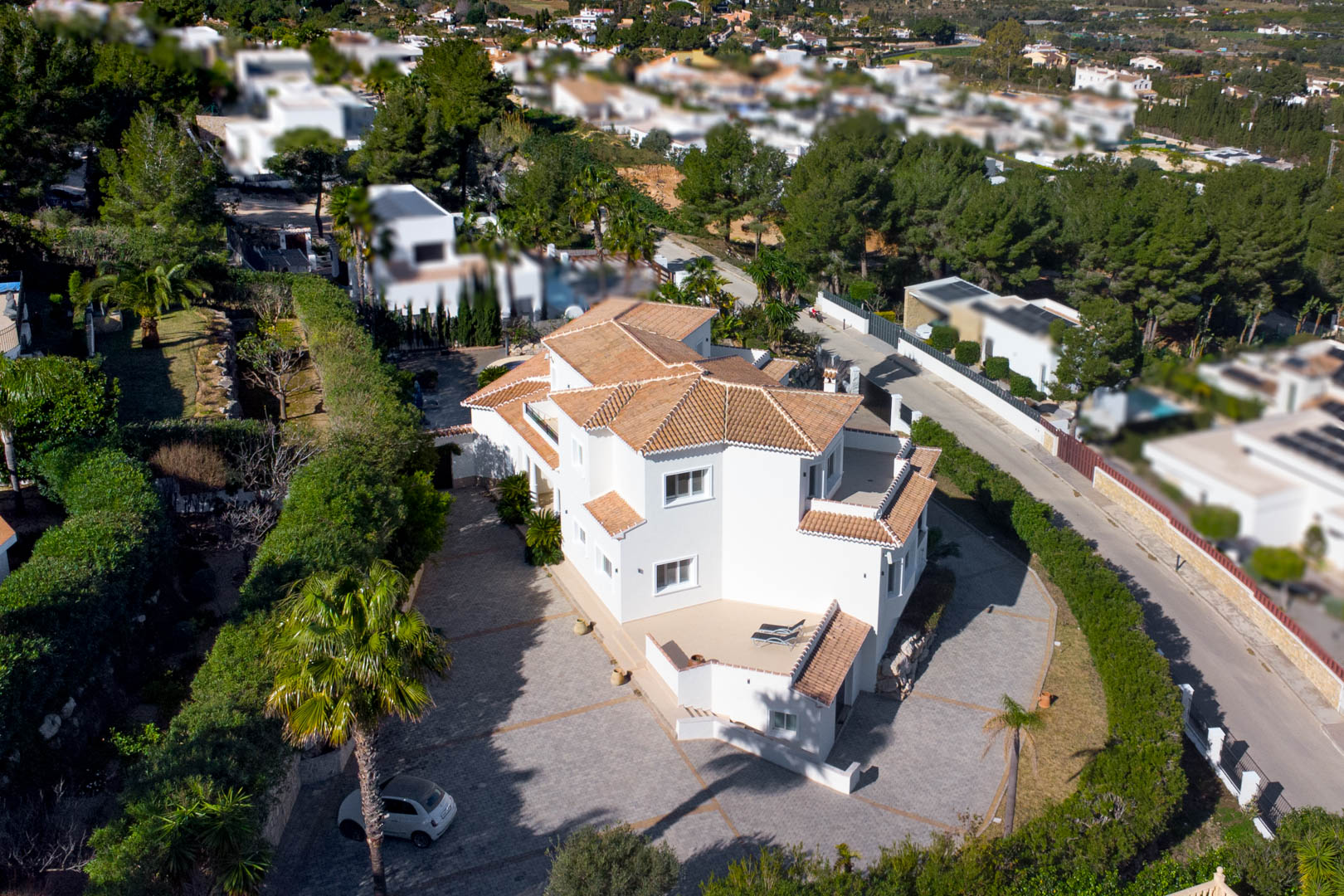Vista aérea de la villa blanca con tejado de teja, destacando su amplia zona de entrada pavimentada y las terrazas privadas rodeadas de vegetación mediterránea.