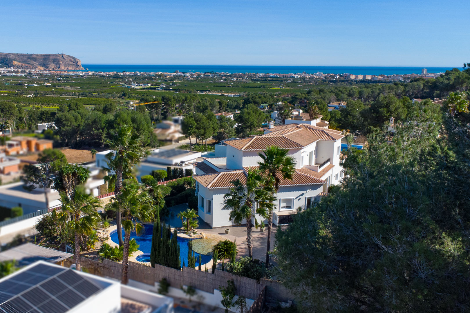 Vista aérea de la villa blanca con tejados de teja, destacando la piscina con jacuzzi rodeada de palmeras y las vistas despejadas hacia el mar Mediterráneo.