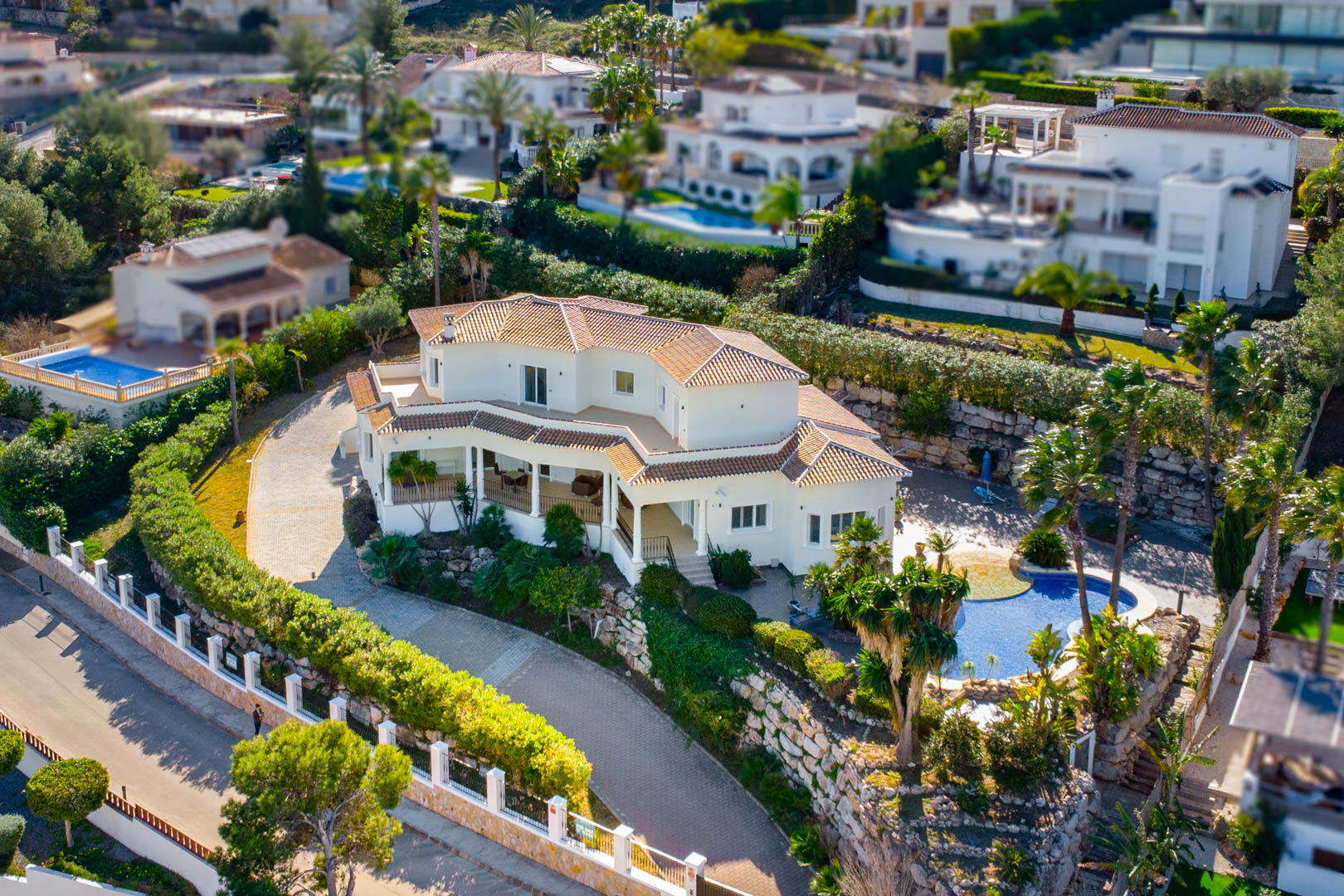 Vista aérea de la villa con fachada blanca y tejado de teja cerámica, destacando su amplia parcela con piscina, jardines mediterráneos y un camino de entrada pavimentado.