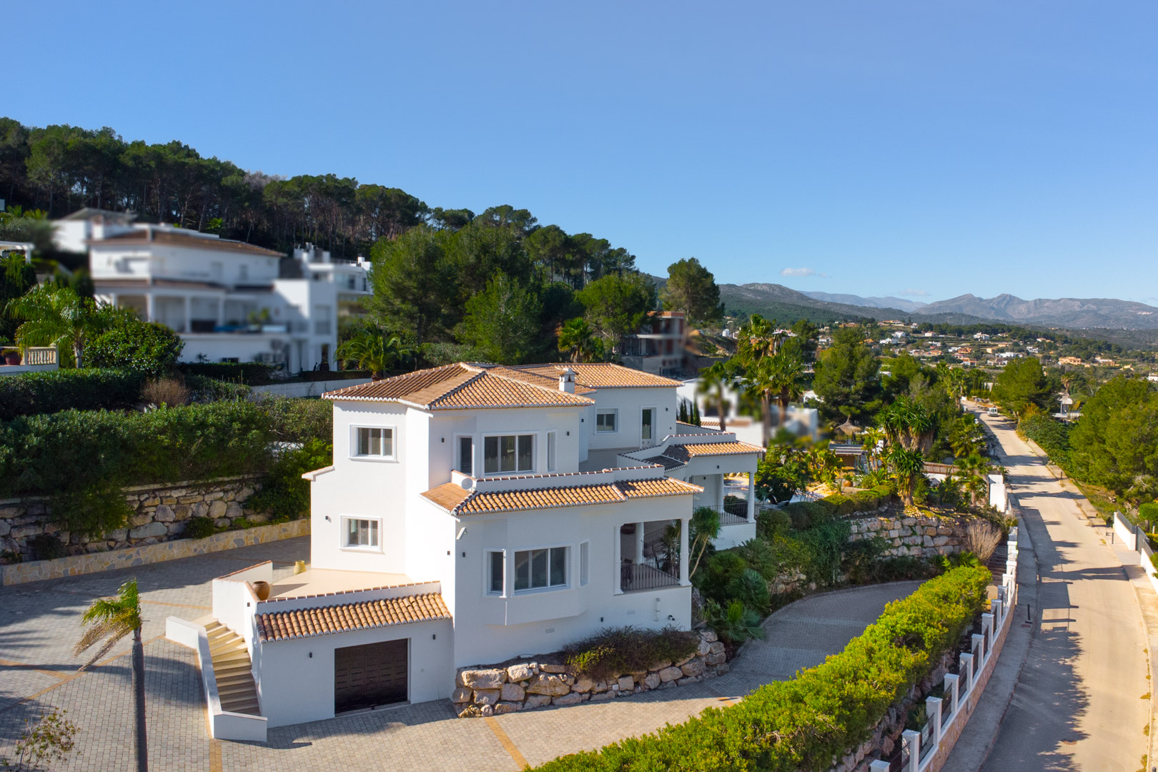 Vista exterior de la villa con fachada blanca y tejados de teja cerámica, destacando sus múltiples niveles, terrazas privadas y el acceso al garaje integrado.