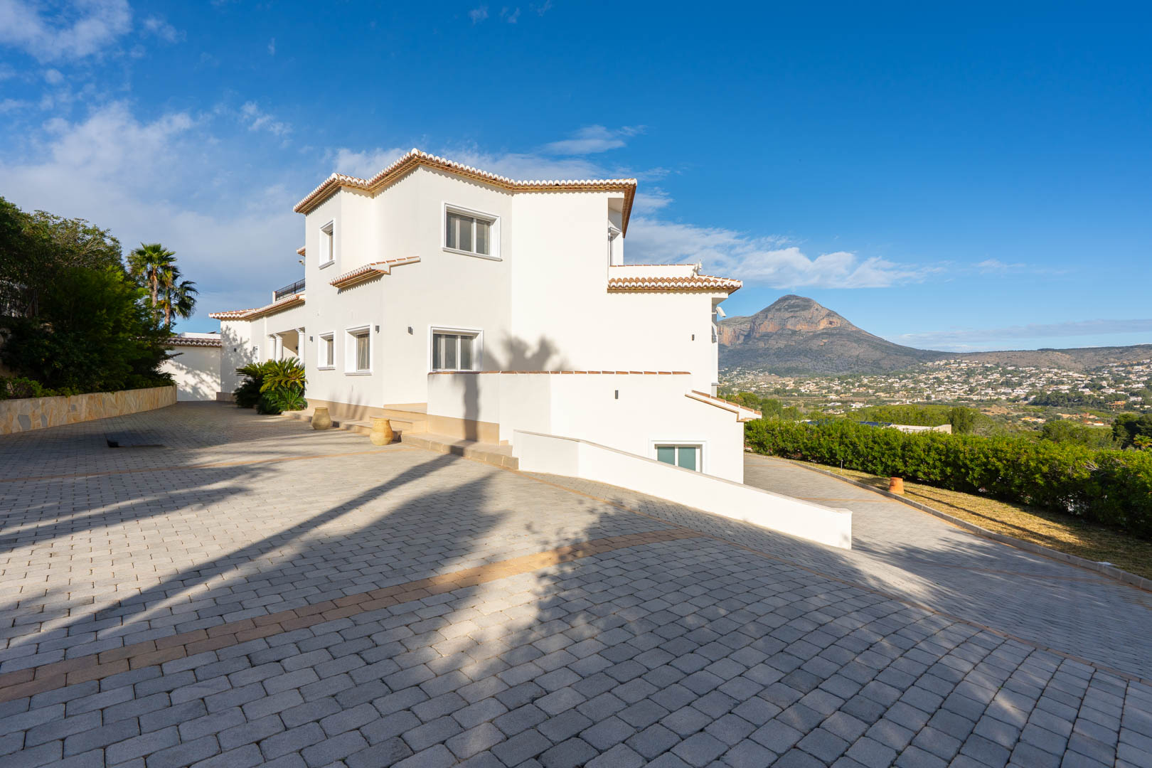Vista exterior de la villa con fachada blanca y tejados de teja, destacando el amplio acceso pavimentado y las vistas despejadas hacia el macizo del Montgó.