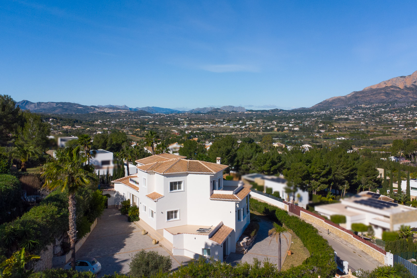 Vista exterior elevada de la villa blanca con tejados de teja, destacando su amplia entrada pavimentada y las vistas panorámicas hacia el valle y el Montgó.