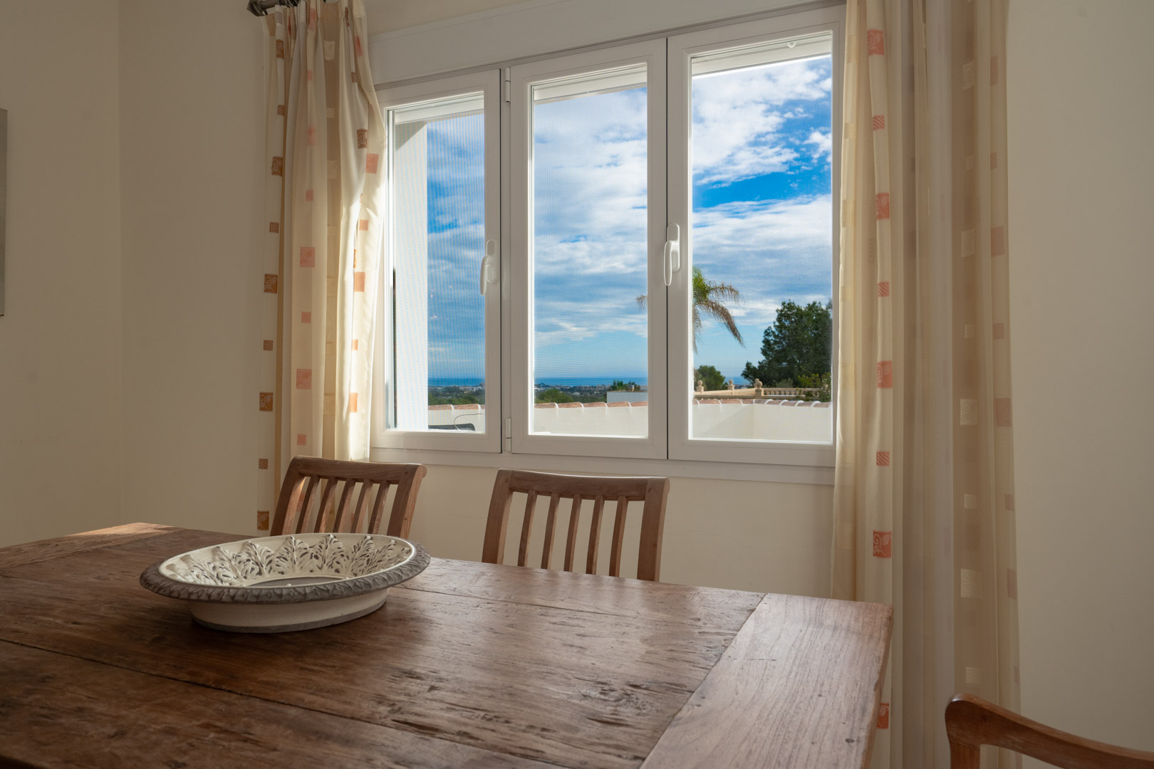 Zona de comedor con mesa de madera rústica y vistas al mar Mediterráneo a través de un ventanal blanco de tres hojas que aporta abundante luz natural.