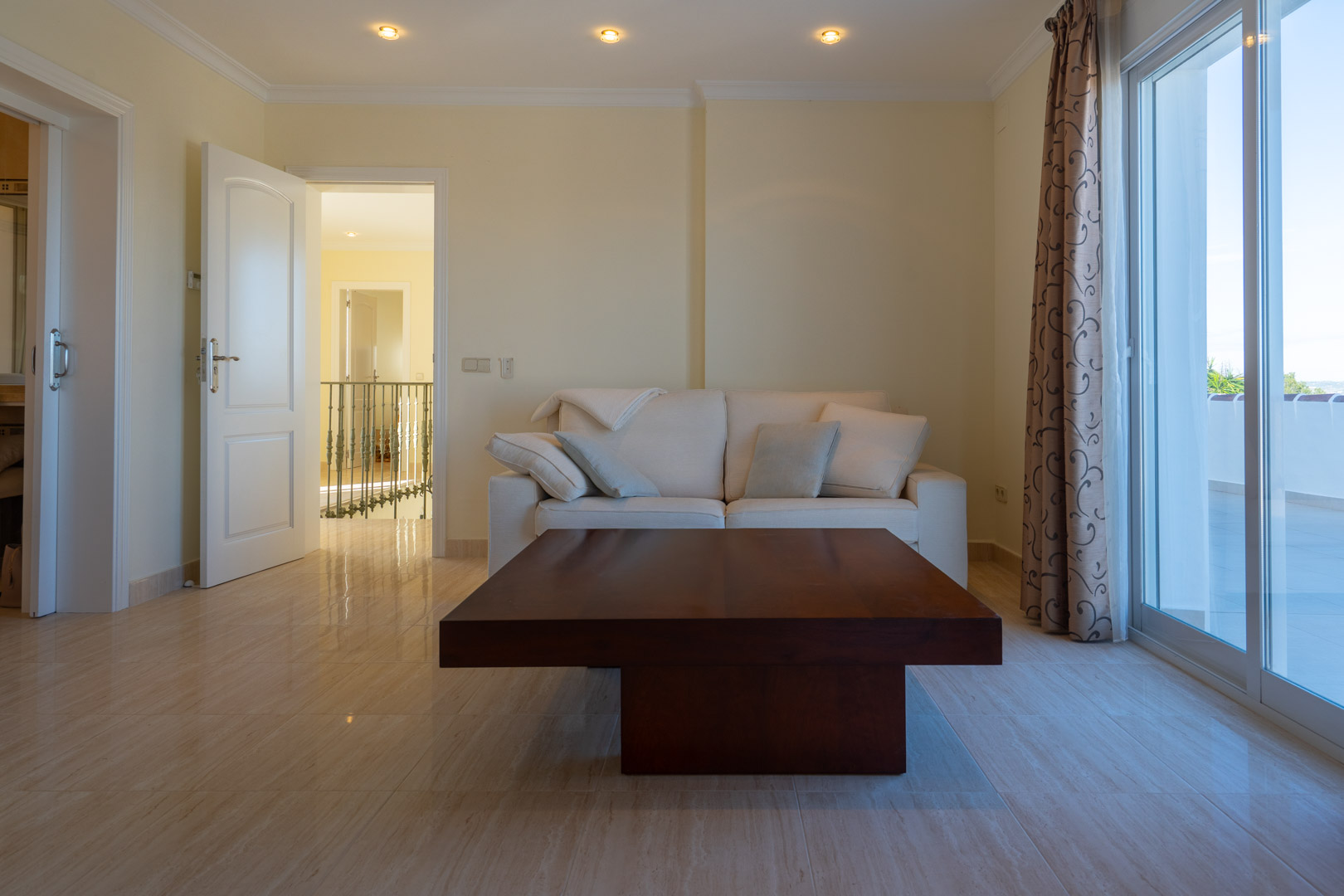 Zona de estar con suelos de mármol pulido, sofá blanco y mesa de madera noble, con acceso directo a la terraza y vistas al rellano de la escalera.