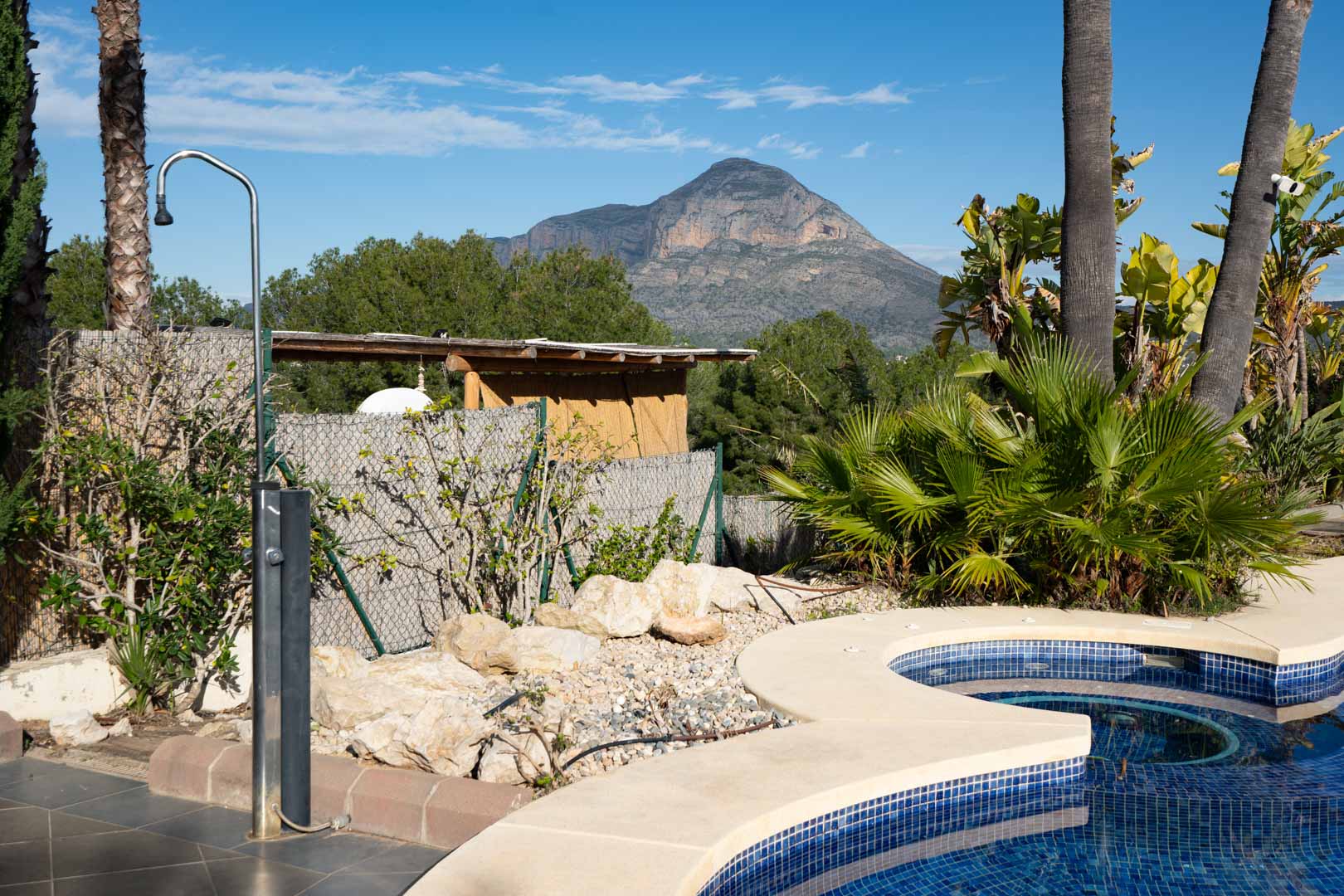 Zona de piscina con ducha exterior de acero inoxidable, rodeada de vegetación tropical y con vistas directas a la montaña del Montgó.