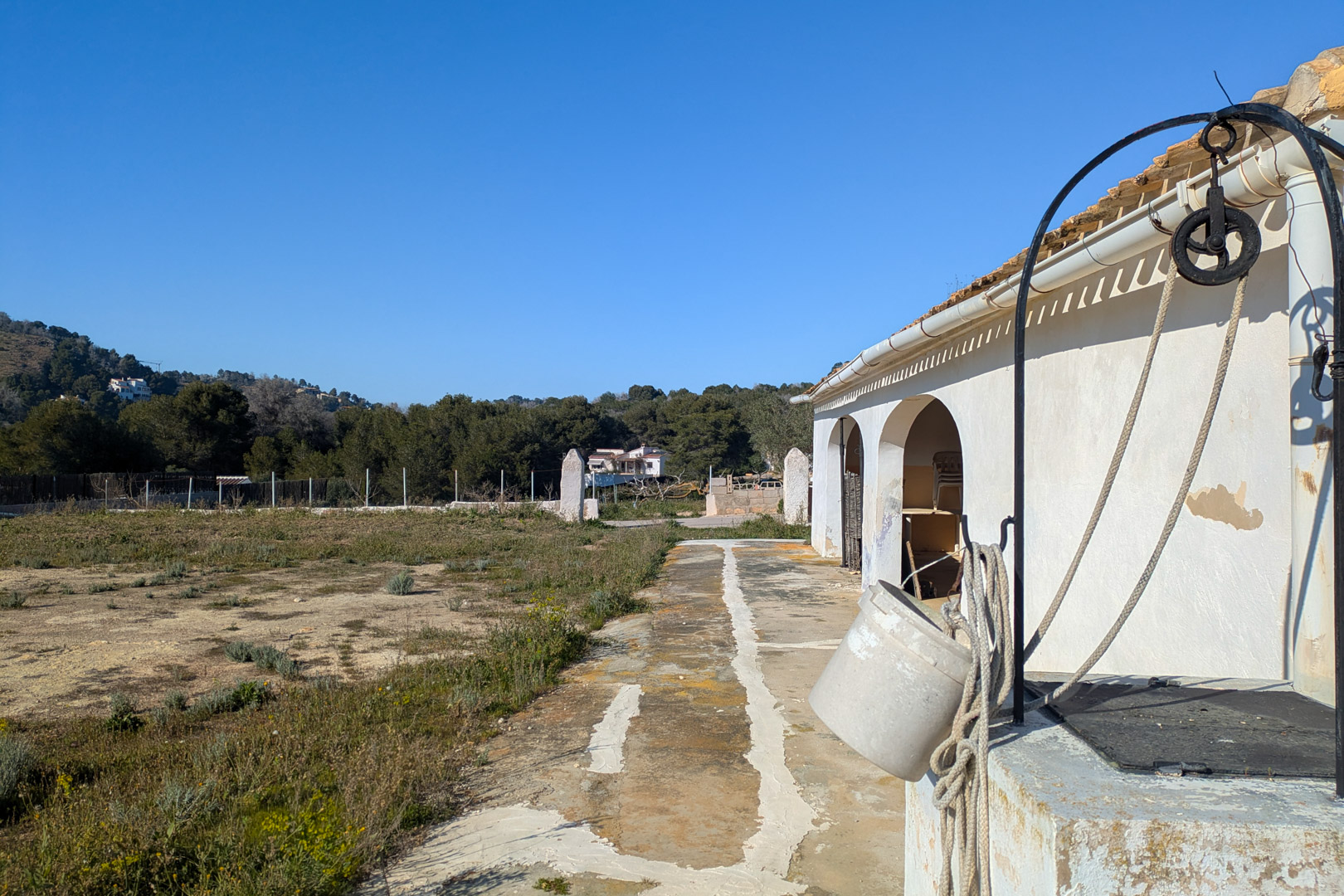 Exterior de la finca con pozo tradicional y polea, situado junto a la fachada blanca con arcos y una extensa parcela rústica llana rodeada de naturaleza.