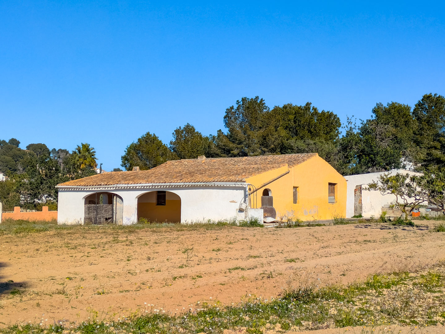 Fachada de la finca tradicional con arcos típicos y tejado de teja cerámica, situada en una amplia parcela rústica rodeada de vegetación mediterránea.