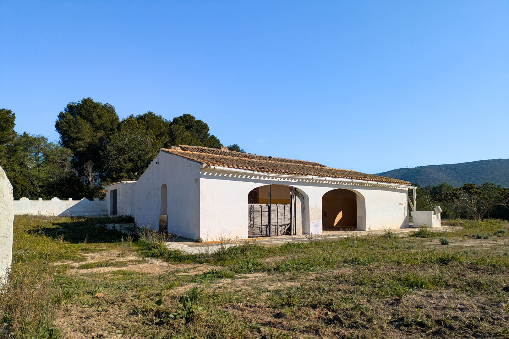 Fachada de la finca tradicional con naya de arcos y tejado de teja árabe, situada en una amplia parcela rústica rodeada de vegetación y pinos.