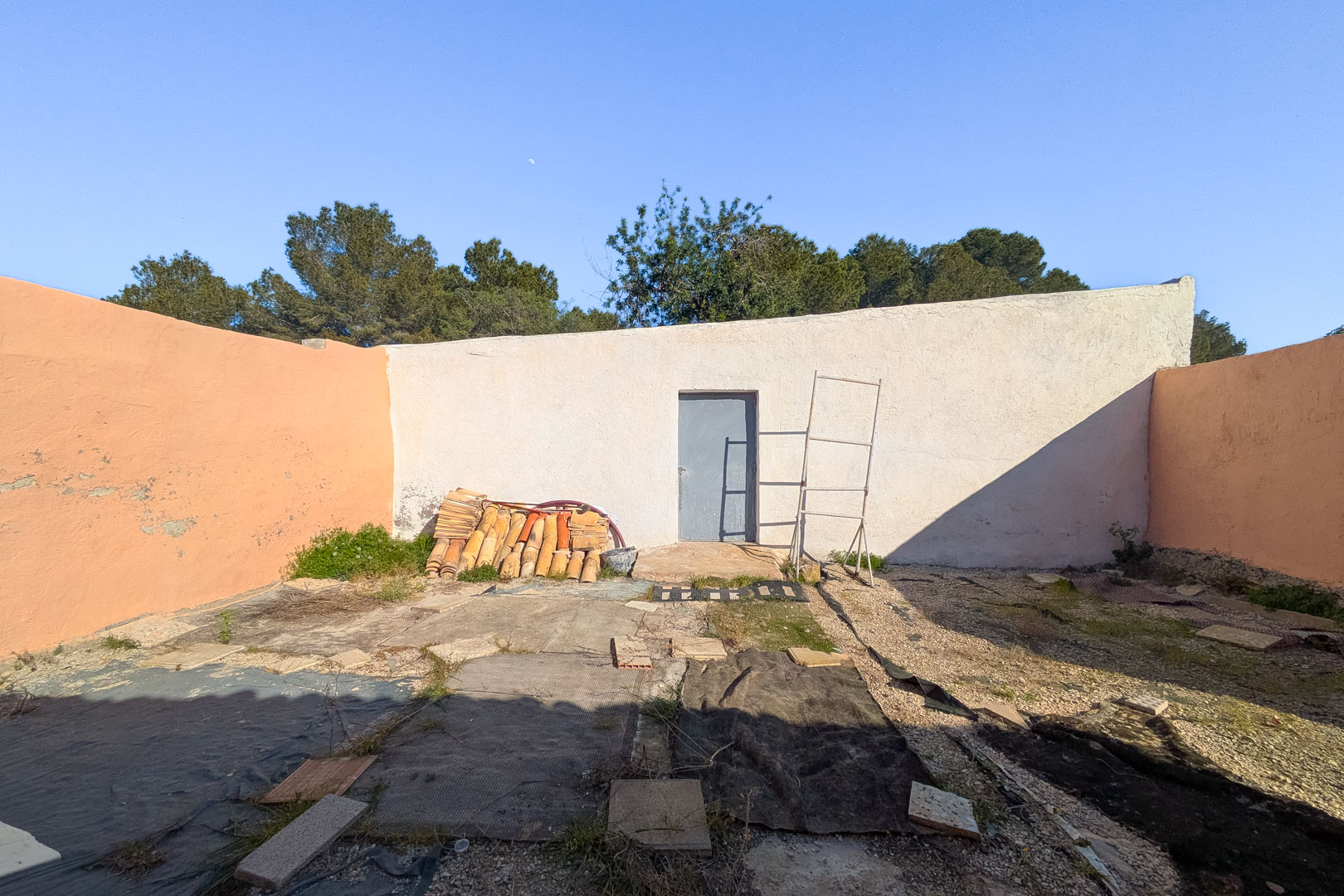 Patio exterior delimitado por muros altos, con una puerta de acceso y materiales de construcción como tejas cerámicas apiladas sobre terreno rústico.