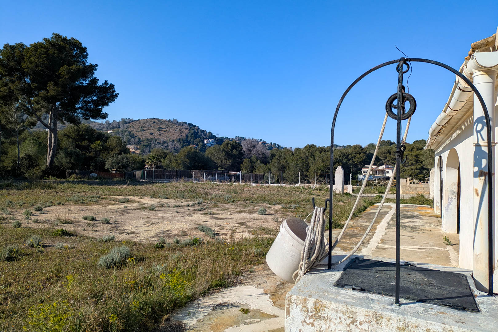 Pozo tradicional con arco de hierro y polea situado en la amplia parcela rústica, ofreciendo vistas despejadas a las colinas y al entorno natural de Jávea.