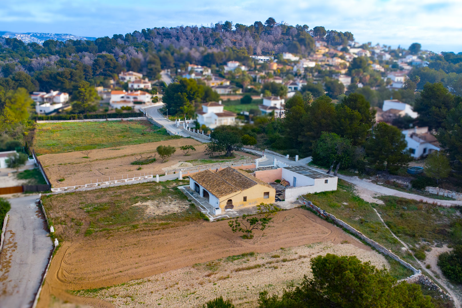 Vista aérea de la finca tradicional con tejado de teja cerámica y fachada ocre, situada en una extensa parcela rústica rodeada de vegetación y chalets.