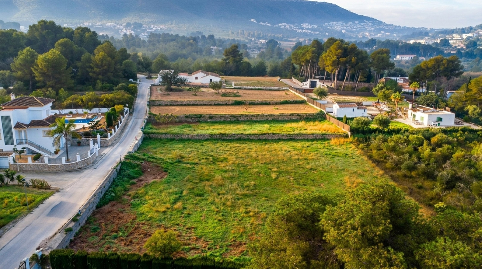 Vista aérea de la parcela rústica organizada en bancales de piedra, rodeada de vegetación mediterránea y con las montañas de Jávea al fondo.