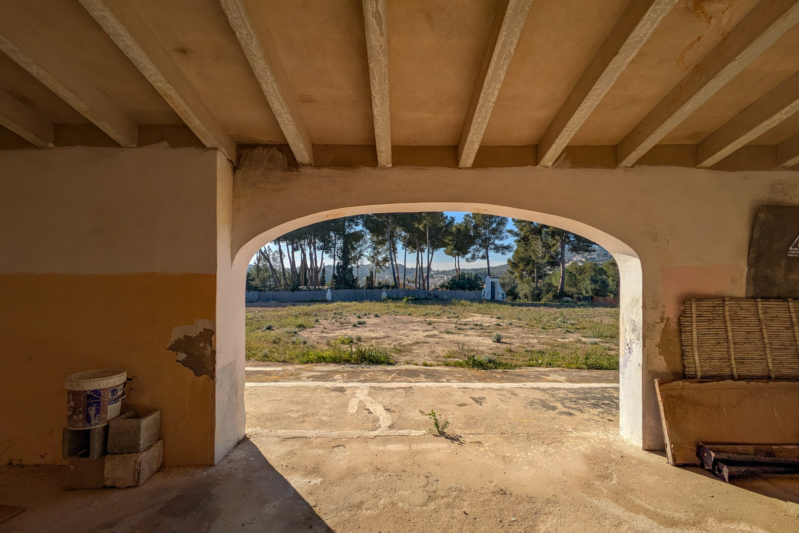 Vista desde el interior de un porche con arcos, mostrando techos de vigas y una amplia parcela rústica con pinos maduros al fondo.