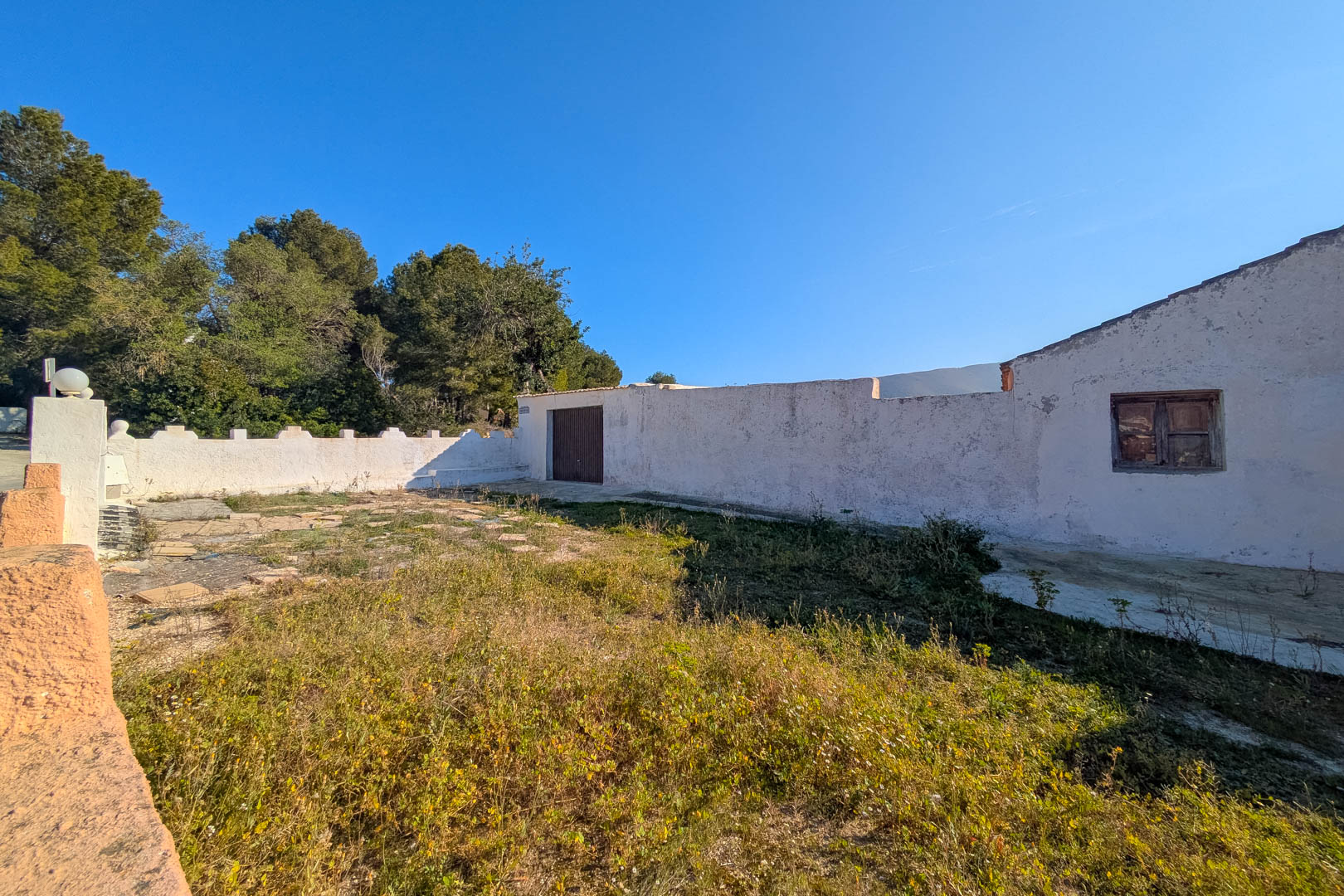 Vista exterior de la fachada blanca con carpintería de madera original y patio rústico delimitado por muros, rodeado de vegetación mediterránea y pinos.