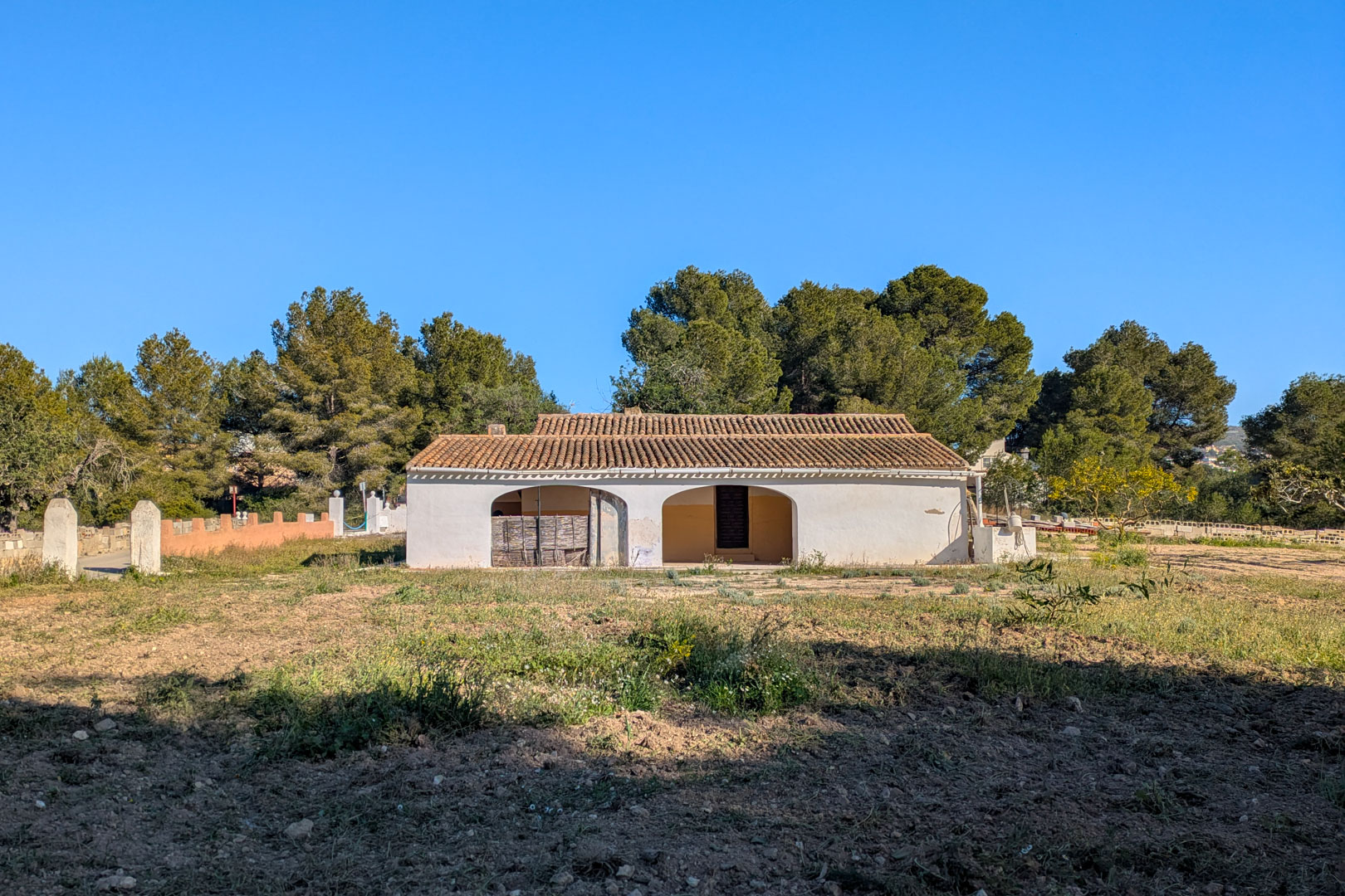Vista exterior de la finca tradicional con fachada blanca, porche de arcos y tejado de teja árabe, rodeada de terreno rústico y pinos bajo un cielo despejado.