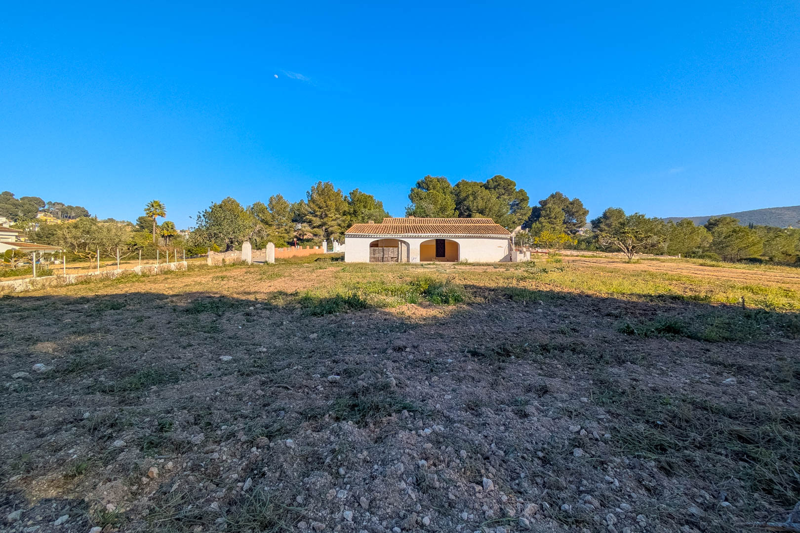 Vista exterior de la finca tradicional con porche de arcos y tejado de teja cerámica, situada en una extensa parcela llana rodeada de pinos.