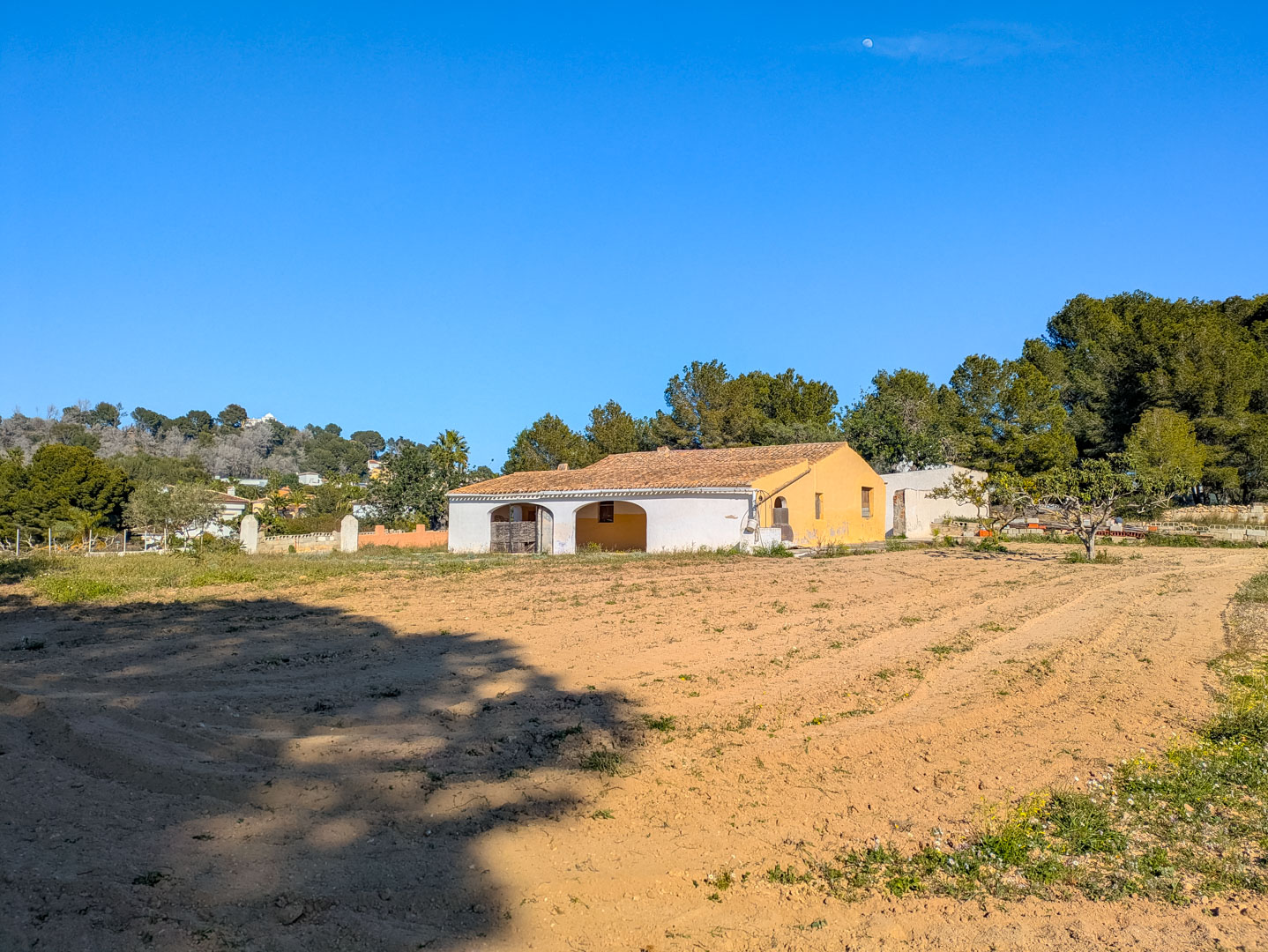 Vista exterior de la finca tradicional con su porche de arcos, tejado de teja árabe y una extensa parcela rústica llana rodeada de pinos mediterráneos.
