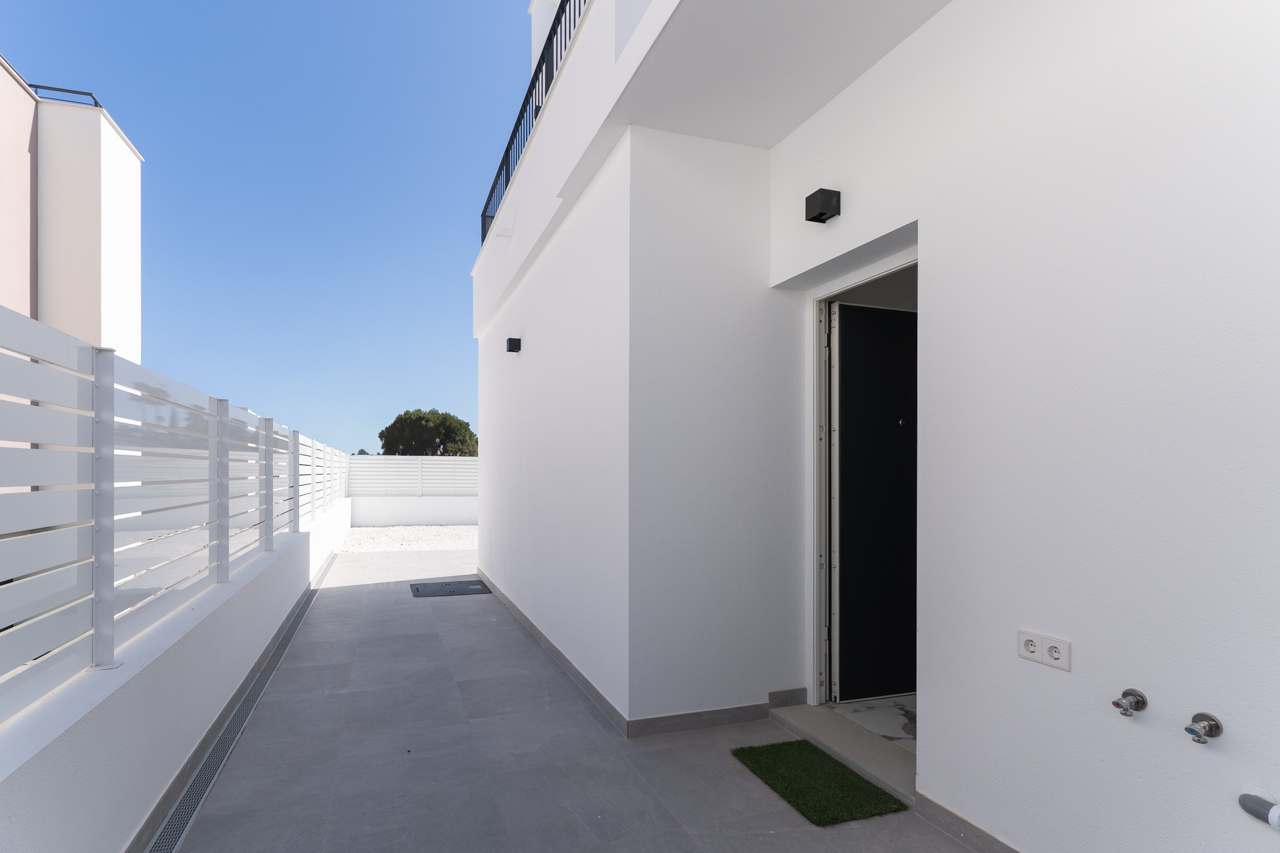 Acceso principal y pasillo exterior con pavimentos cerámicos grises, paredes blancas lisas y vallado perimetral de lamas, destacando la arquitectura moderna y lineal.