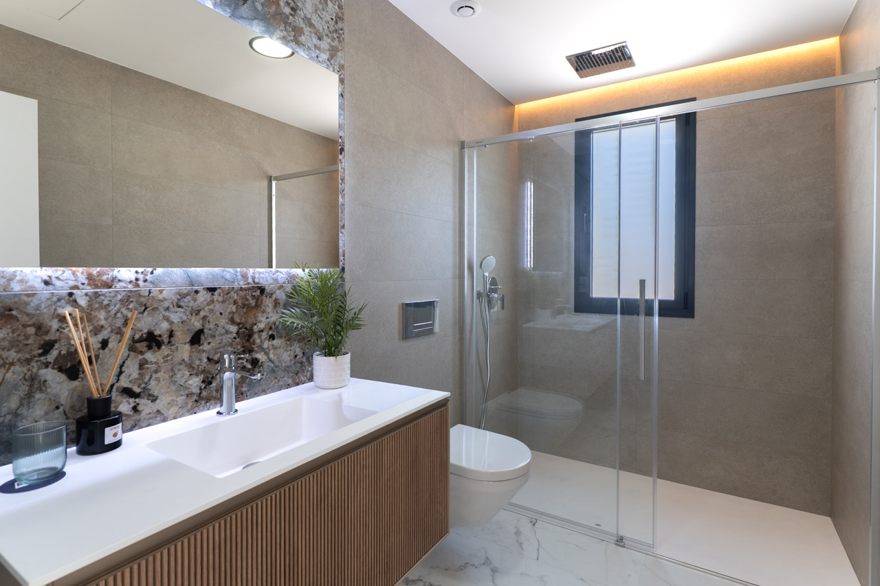Baño moderno con mueble de madera alistonada, salpicadero de mármol y ducha italiana con mampara de cristal e iluminación LED indirecta.