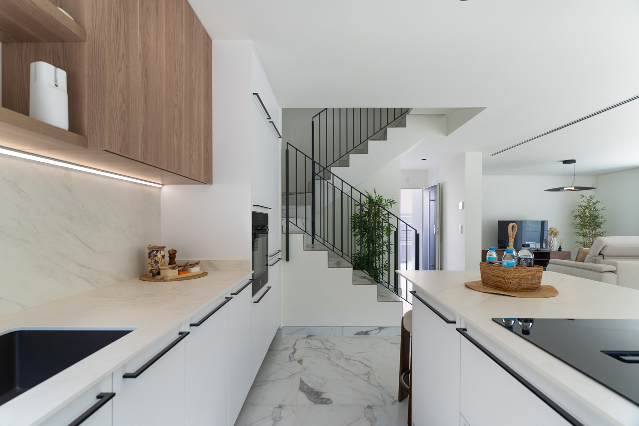 Cocina de concepto abierto con mobiliario blanco, encimeras marmoleadas y armarios superiores de madera con iluminación LED. Al fondo se aprecia la escalera de diseño y el salón.
