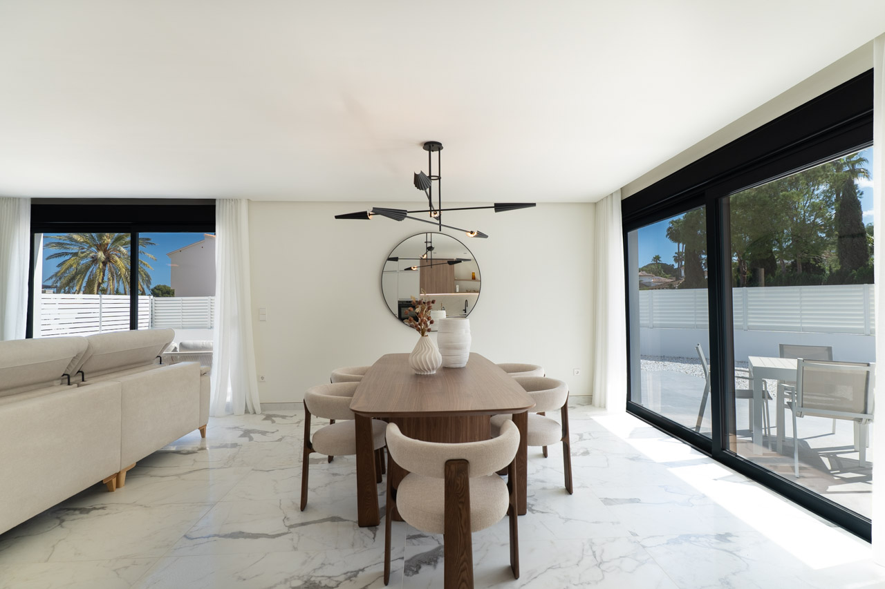 Comedor de concepto abierto con mesa de madera, sillas tapizadas y suelos de mármol. Grandes ventanales conectan el interior con la terraza exterior.