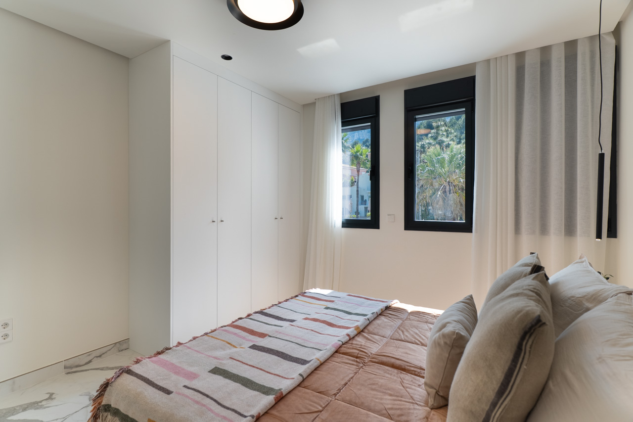 Dormitorio luminoso con armarios empotrados blancos, suelo de mármol y ventanas con vistas al entorno natural de Dénia.