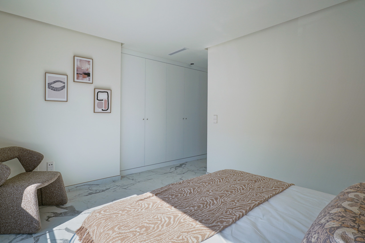 Dormitorio luminoso con armarios empotrados blancos, suelos de mármol veteado y un rincón de lectura con sillón de diseño y cuadros minimalistas.