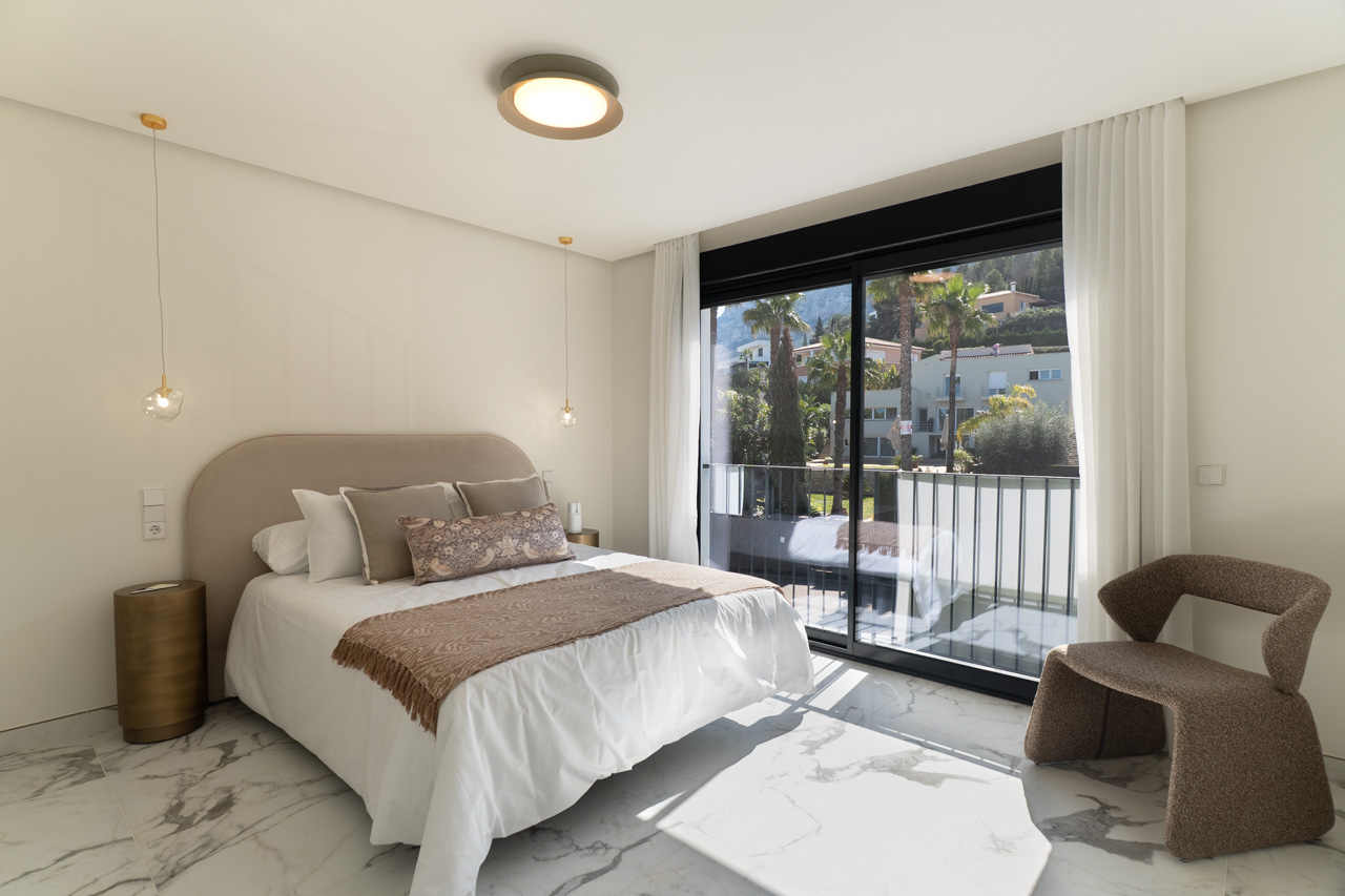Dormitorio principal con suelos de gres porcelánico efecto mármol y ventanal de suelo a techo con acceso directo al balcón y vistas al entorno natural.