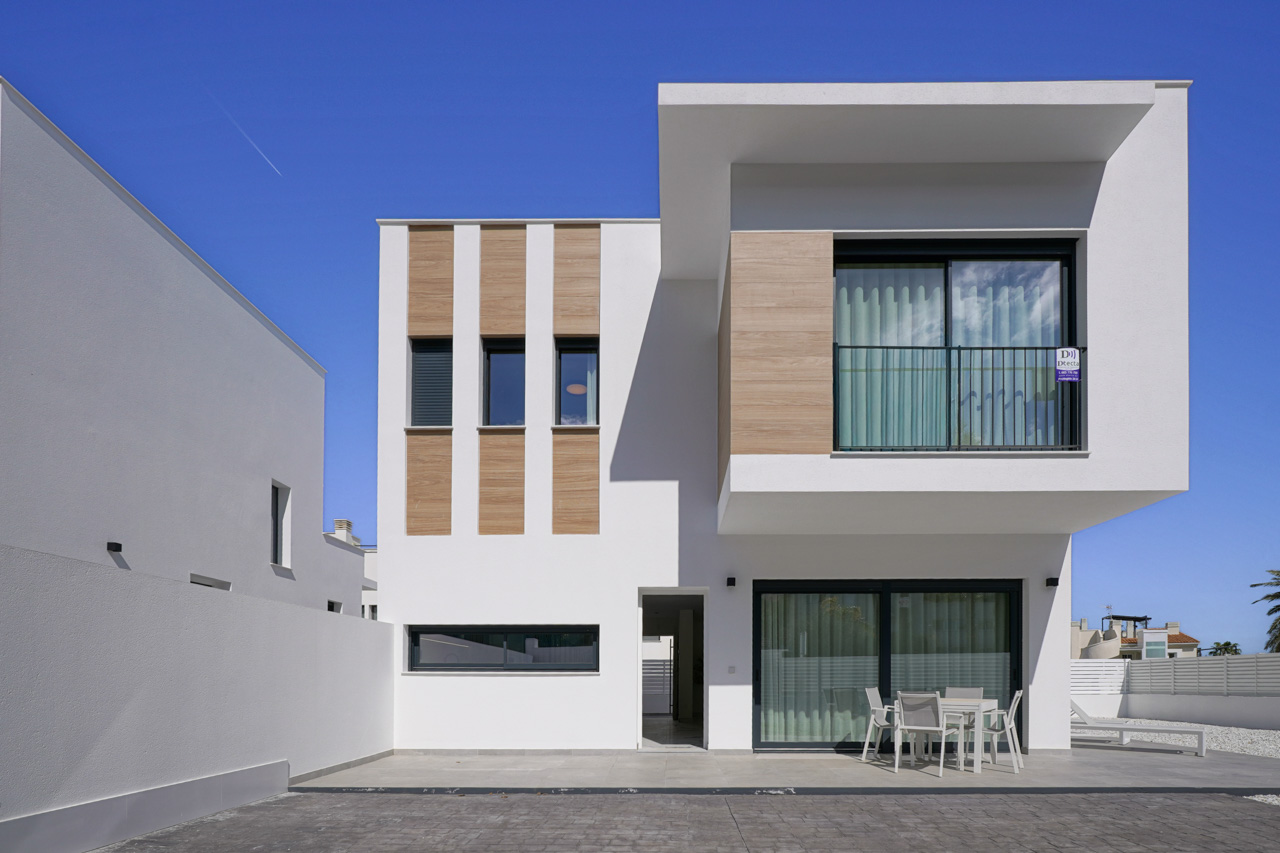 Fachada principal de diseño contemporáneo con acabados en blanco y paneles de efecto madera, destacando la terraza inferior y el balcón superior con grandes ventanales.