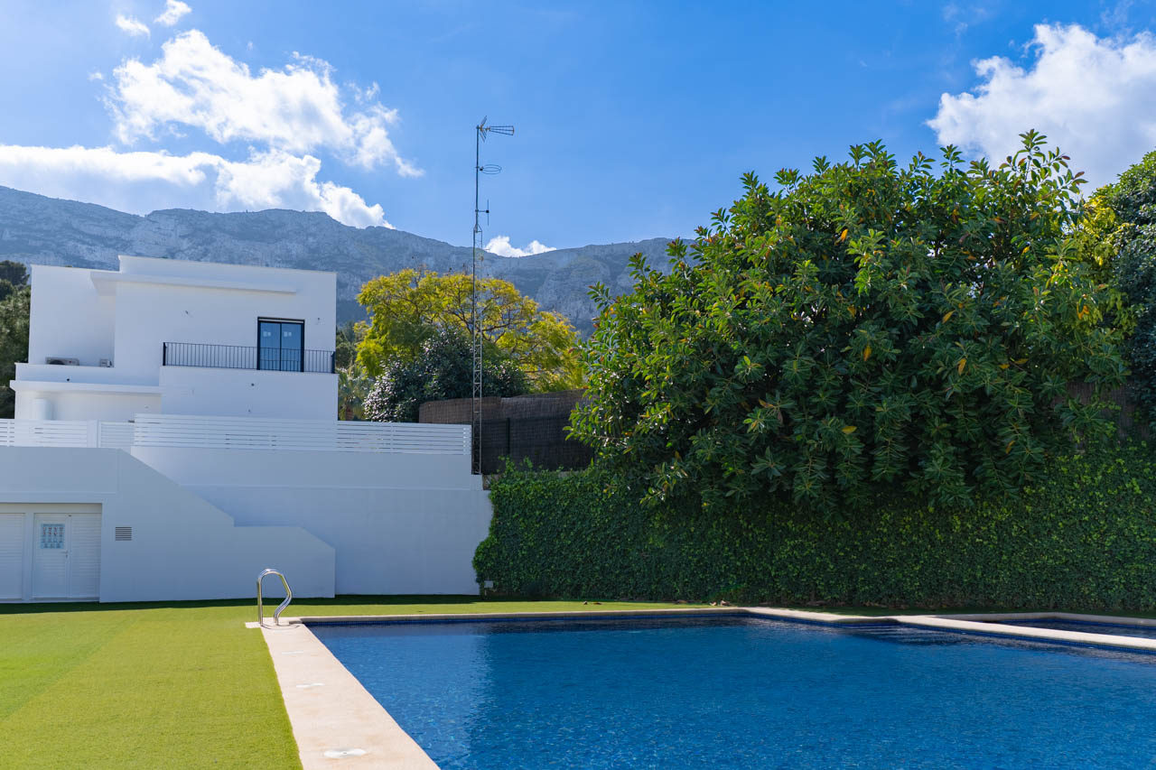 Piscina rectangular con borde de piedra y zona de césped, situada frente a la fachada blanca de estilo minimalista con el macizo del Montgó de fondo.