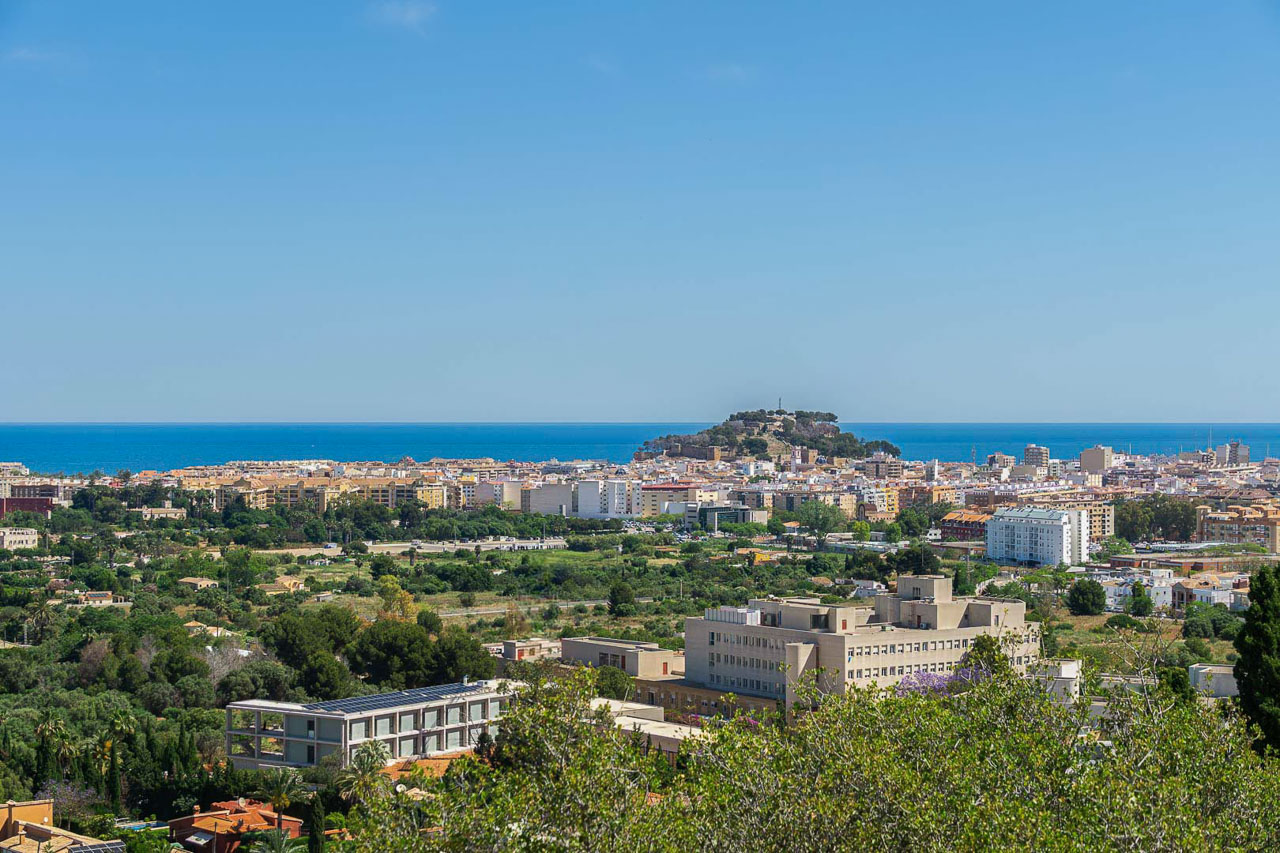 Vistas panorámicas de Dénia con el Castillo y el mar Mediterráneo en el horizonte bajo un cielo despejado. La perspectiva destaca la proximidad al centro urbano y la costa.