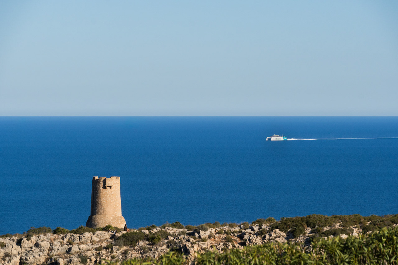 Vistas panorámicas del litoral de Las Rotas en Dénia, destacando la histórica Torre del Gerro y un ferry de Balearia navegando por el mar Mediterráneo.