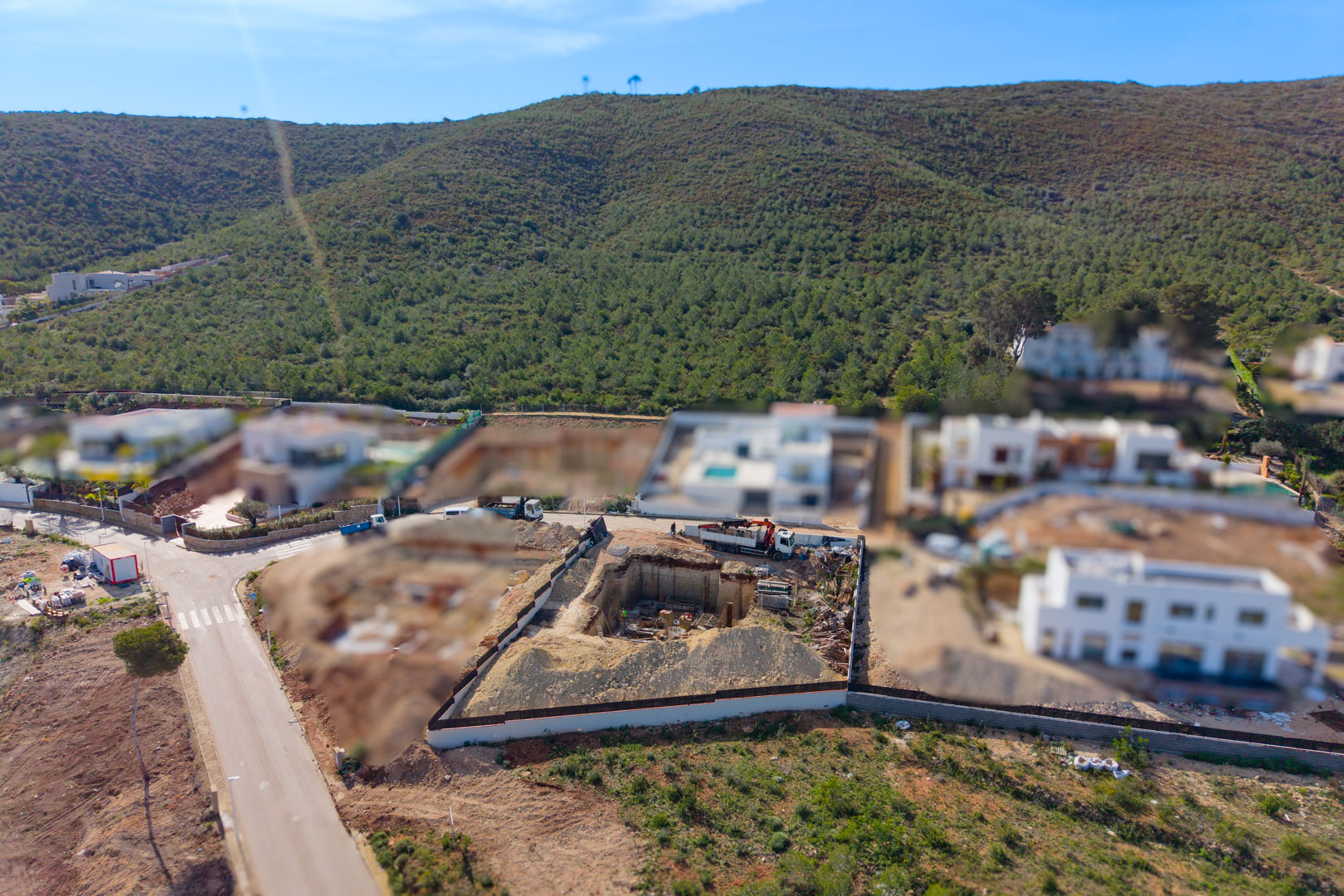 Vista aérea de la parcela en fase de construcción, destacando la excavación profunda para el nivel del sótano y la cimentación en un entorno residencial rodeado de naturaleza.