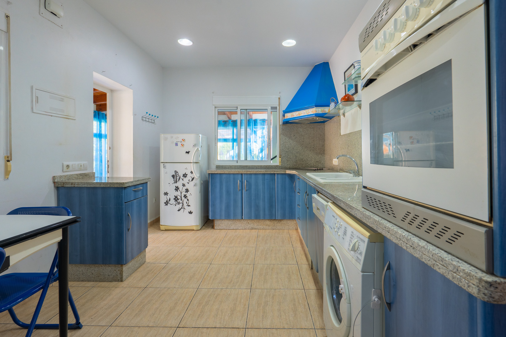 Cocina equipada con armarios azules, encimeras de granito gris y electrodomésticos. Dispone de iluminación empotrada, suelos cerámicos y acceso directo a zonas exteriores.
