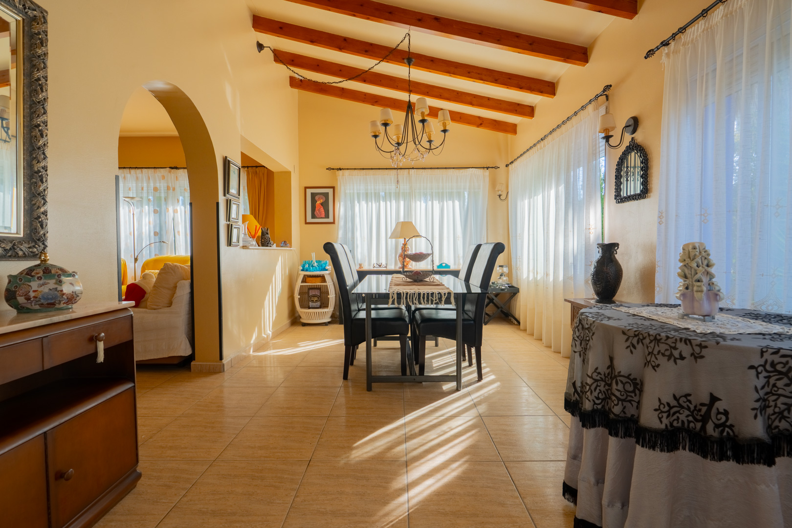 Comedor con techos altos de vigas de madera expuestas, suelos de gres y un arco que comunica con el salón, iluminado por luz natural filtrada.