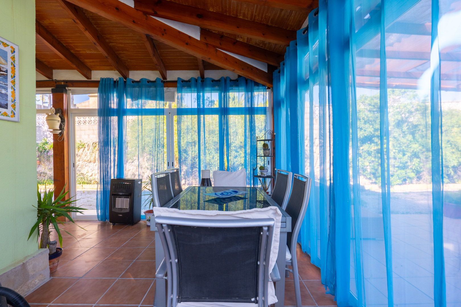 Comedor en naya acristalada con vigas de madera a la vista, suelos de terracota y cortinas azules que filtran la luz natural hacia el jardín.