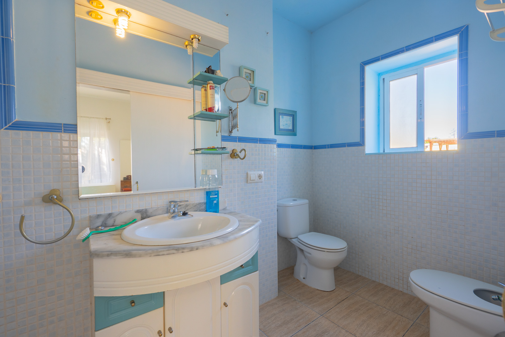 Cuarto de baño con azulejos de mosaico en tonos azules y blancos, encimera de mármol con lavabo integrado y luz natural proveniente de una ventana lateral.