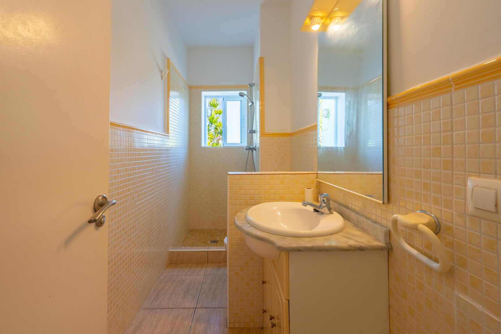 Cuarto de baño con ducha de obra, revestimiento de mosaico en tonos crema y amarillo, encimera de mármol con lavabo encastrado y luz natural.