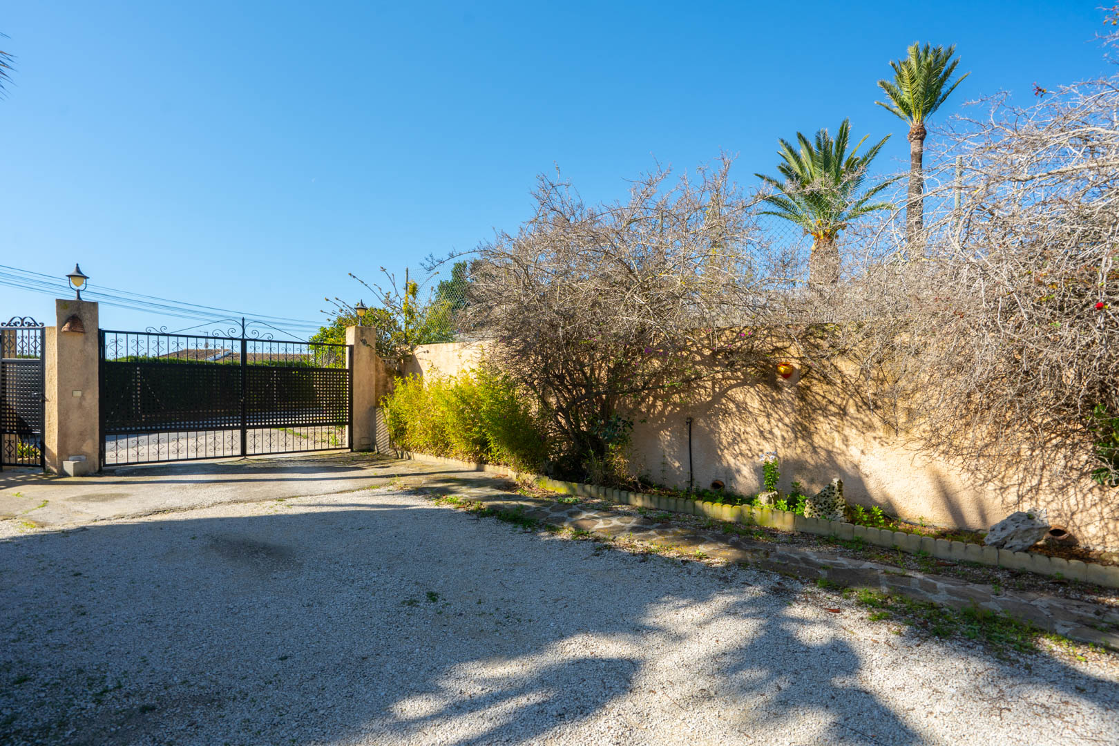 Entrada principal con portón de hierro forjado negro, camino de grava y muro perimetral con vegetación mediterránea y palmeras.