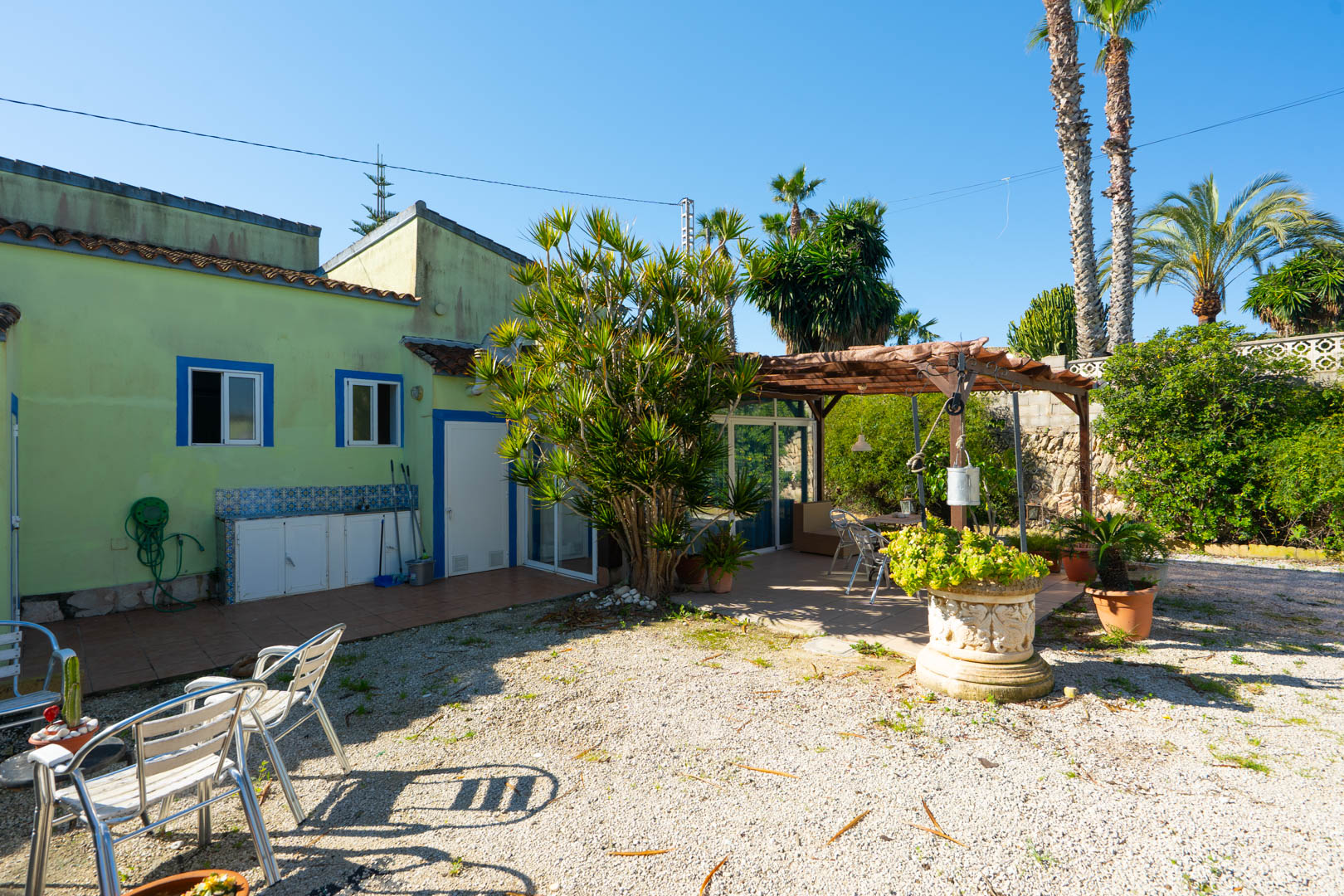 Patio exterior con zona de grava, pérgola de madera y cocina de verano integrada en la fachada verde con detalles azules.