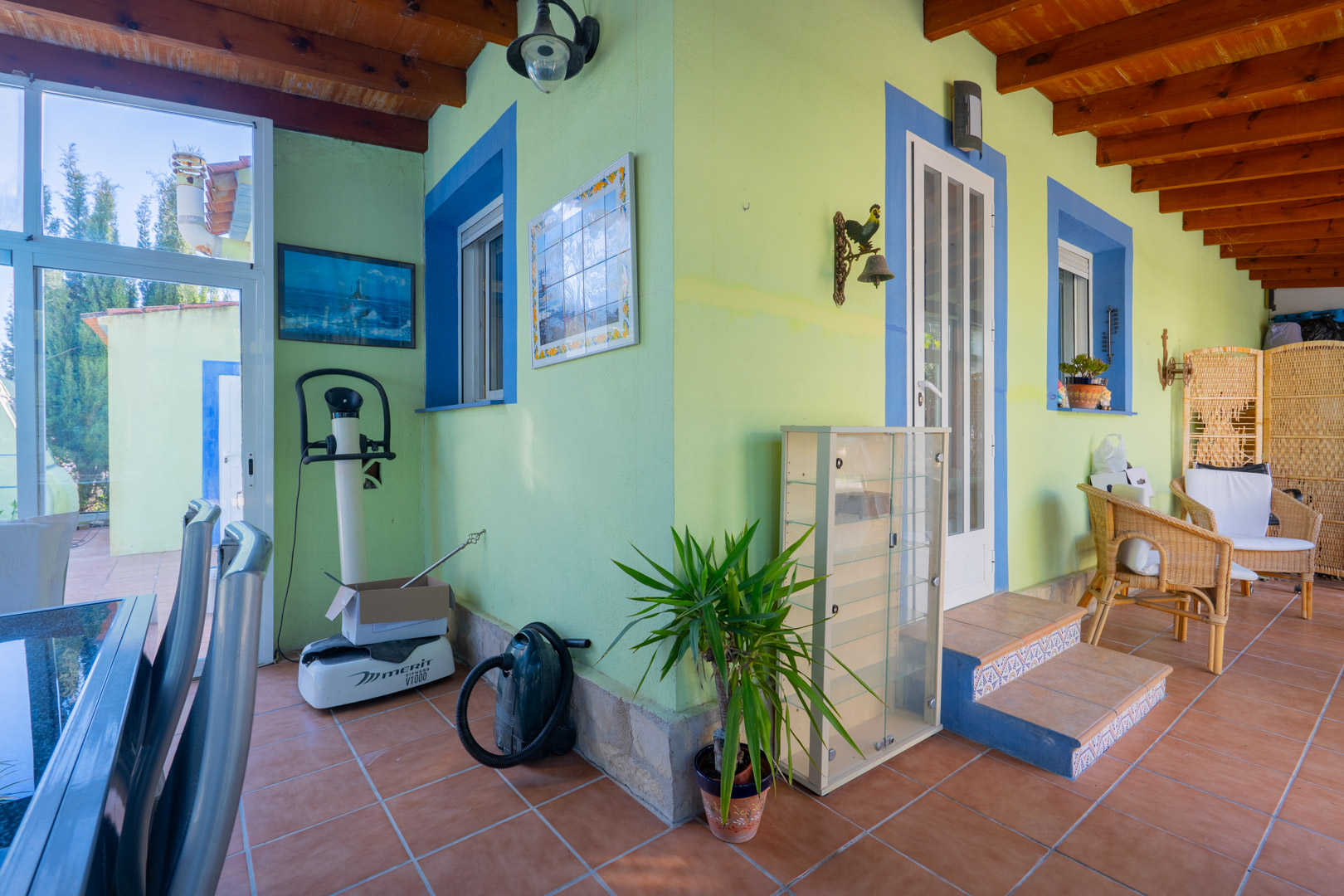 Terraza cubierta con vigas de madera vista, paredes en tonos verde y azul, suelo de gres cerámico y mobiliario de mimbre bajo una naya tradicional.